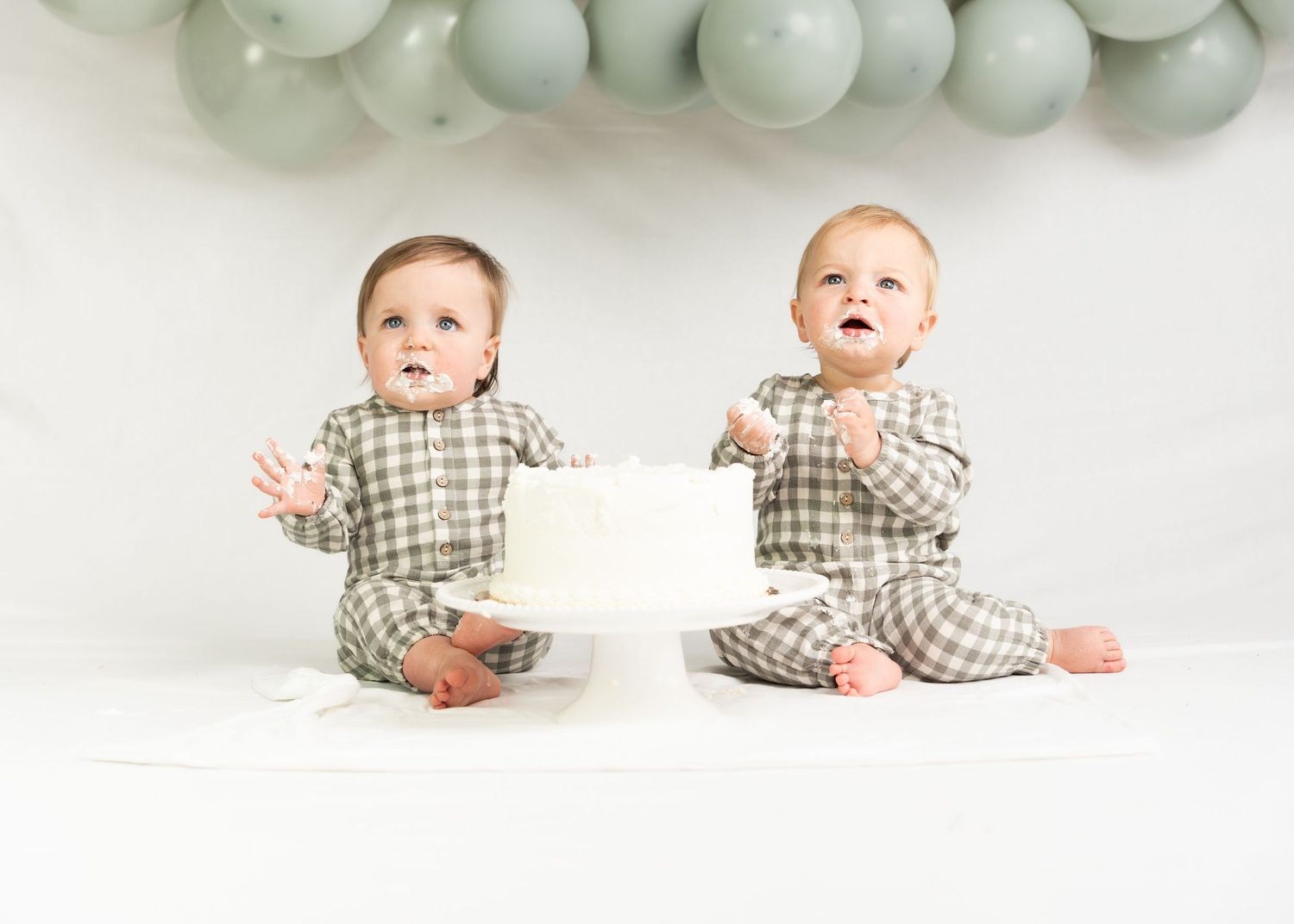 Twin babies in matching green plaid outfits celebrate first birthday with white cake and balloon backdrop.