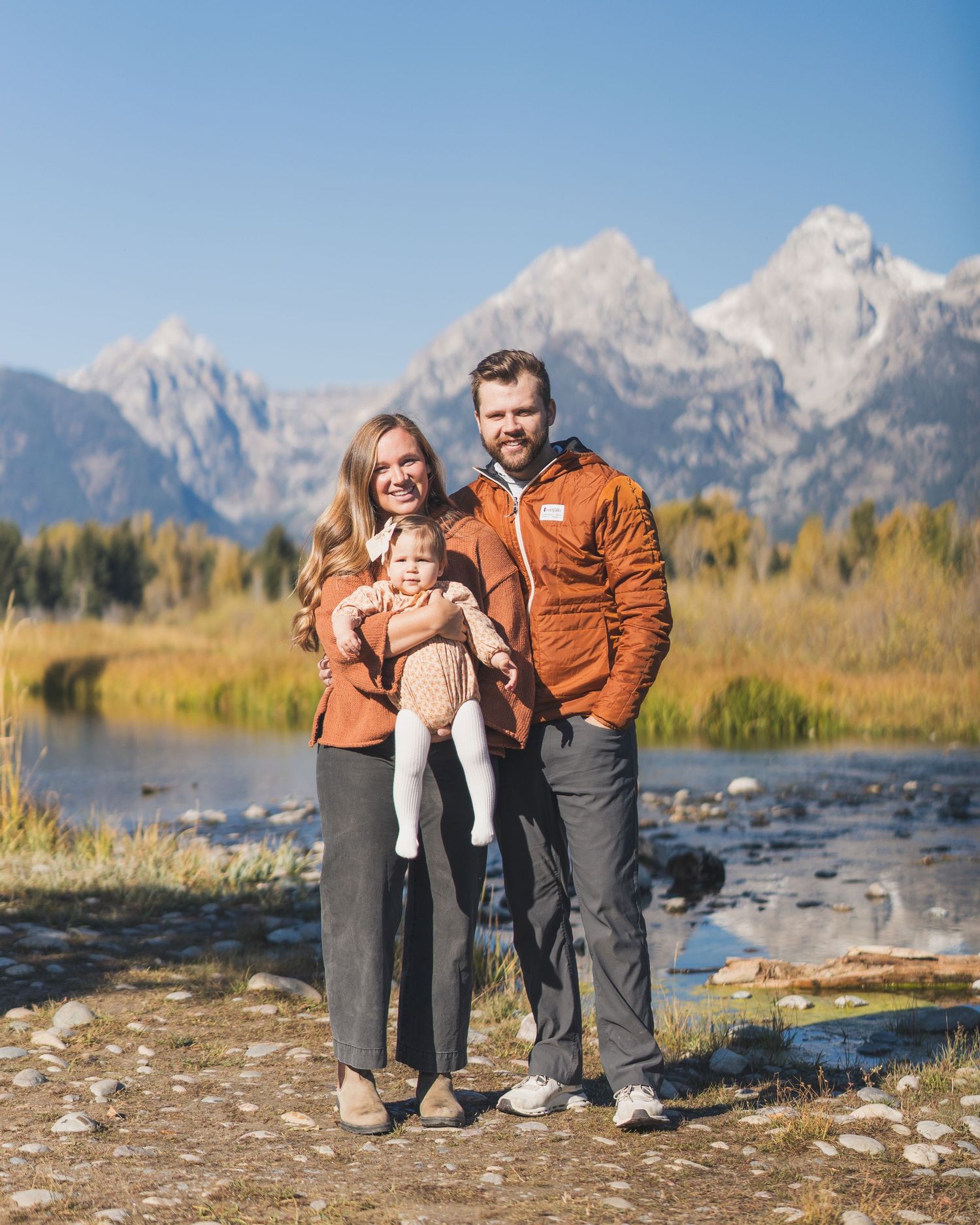 Young family with baby poses before Grand Teton mountains in autumn landscape.