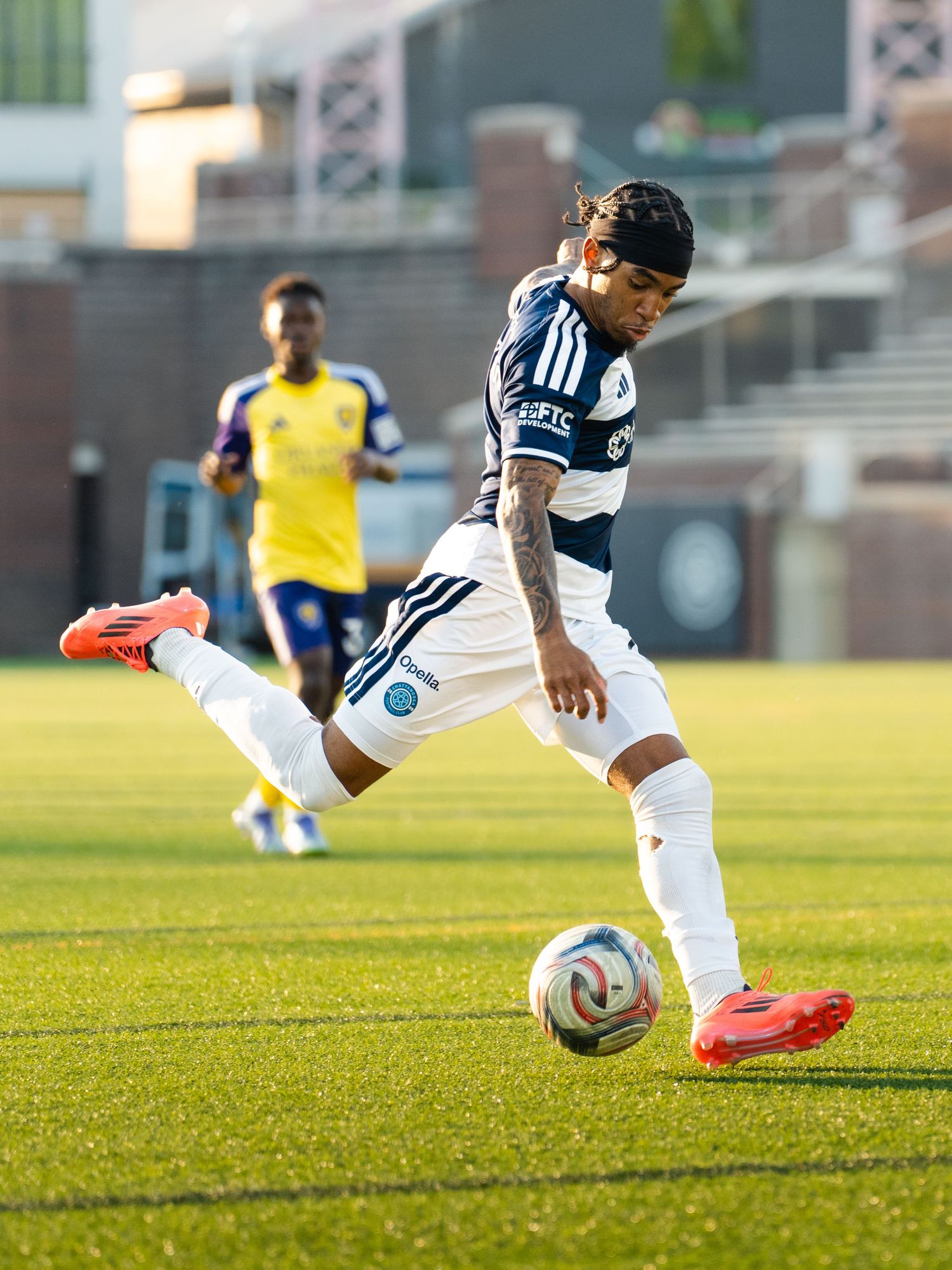 Chattanooga FC soccer player in navy blue kit strikes ball with precision during sunny outdoor MLS Next Pro match.