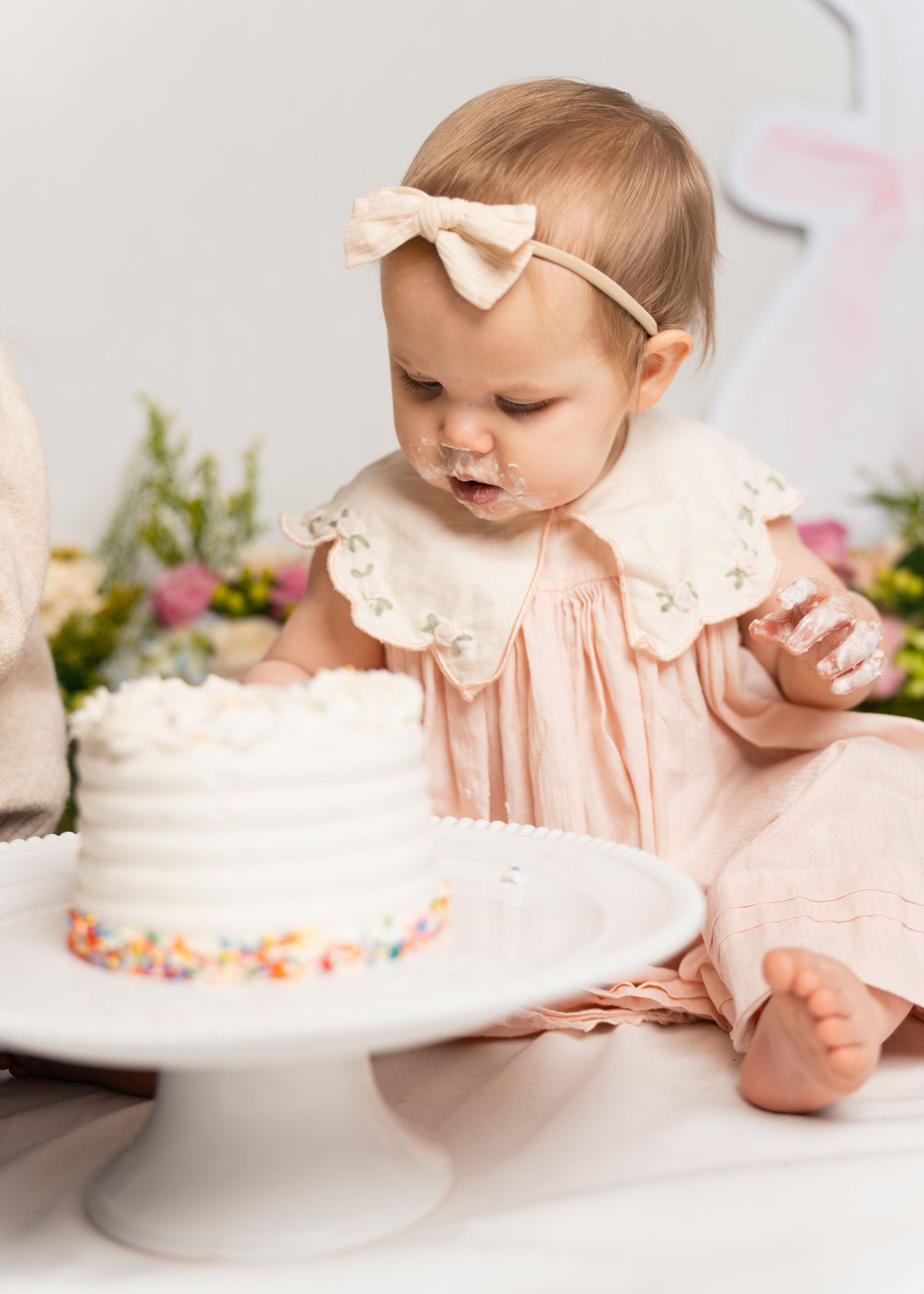 A baby in a pink embroidered dress examines a white frosted birthday cake on a pedestal stand.