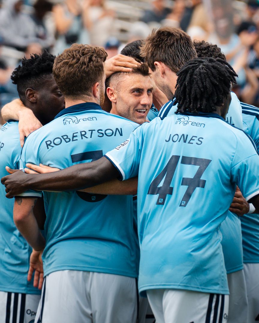 Chattanooga FC Soccer players in light blue jerseys celebrate together on the field, with Robertson and Jones jerseys visible.