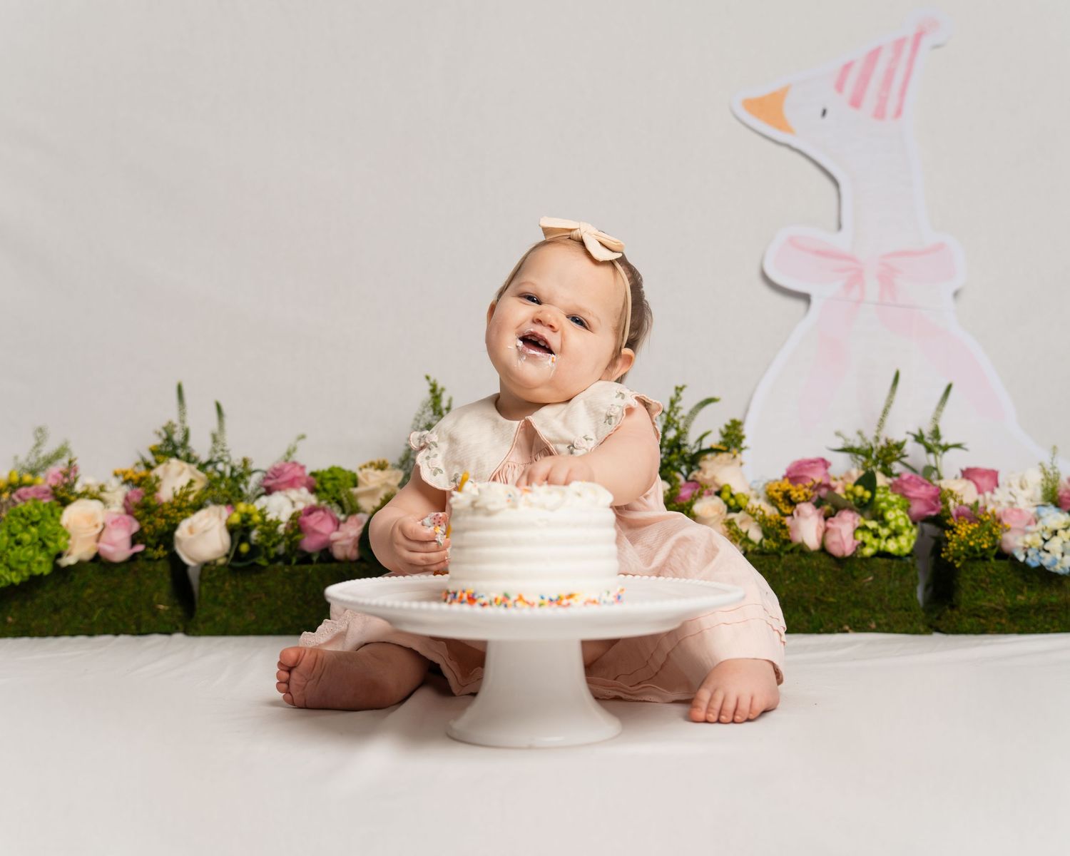 A happy baby in a pink dress gleefully smashes into a white birthday cake on a cake stand.
