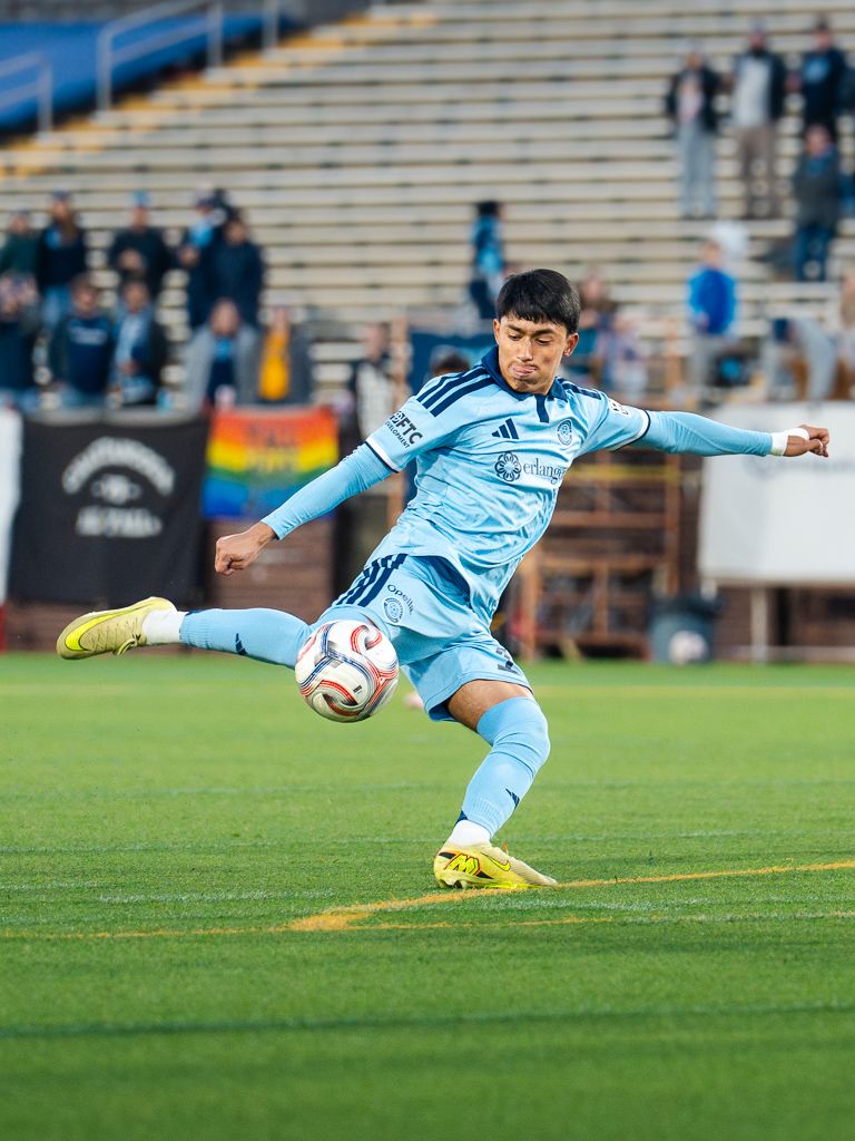 Chattanooga FC Soccer player in light blue kit strikes the ball during a match with fans in the stadium background.