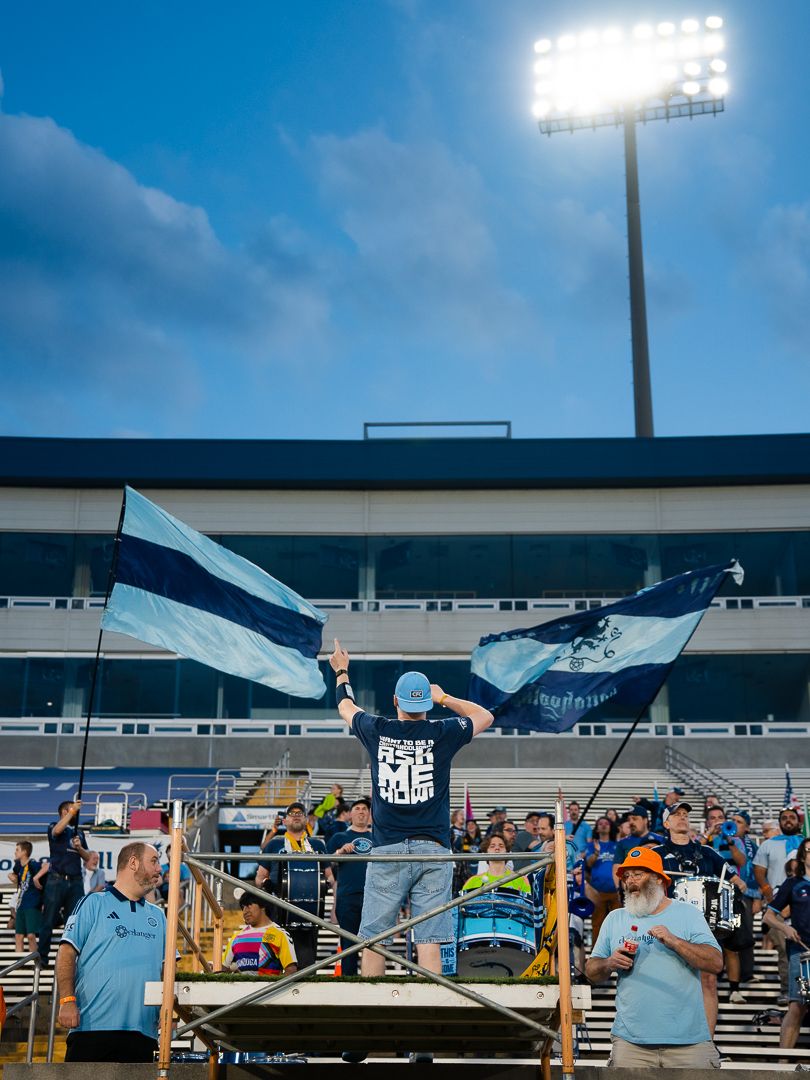 Chattanooga FC Fans (Chattahooligans) waves large blue and white flags in a stadium supporter section under bright floodlights.