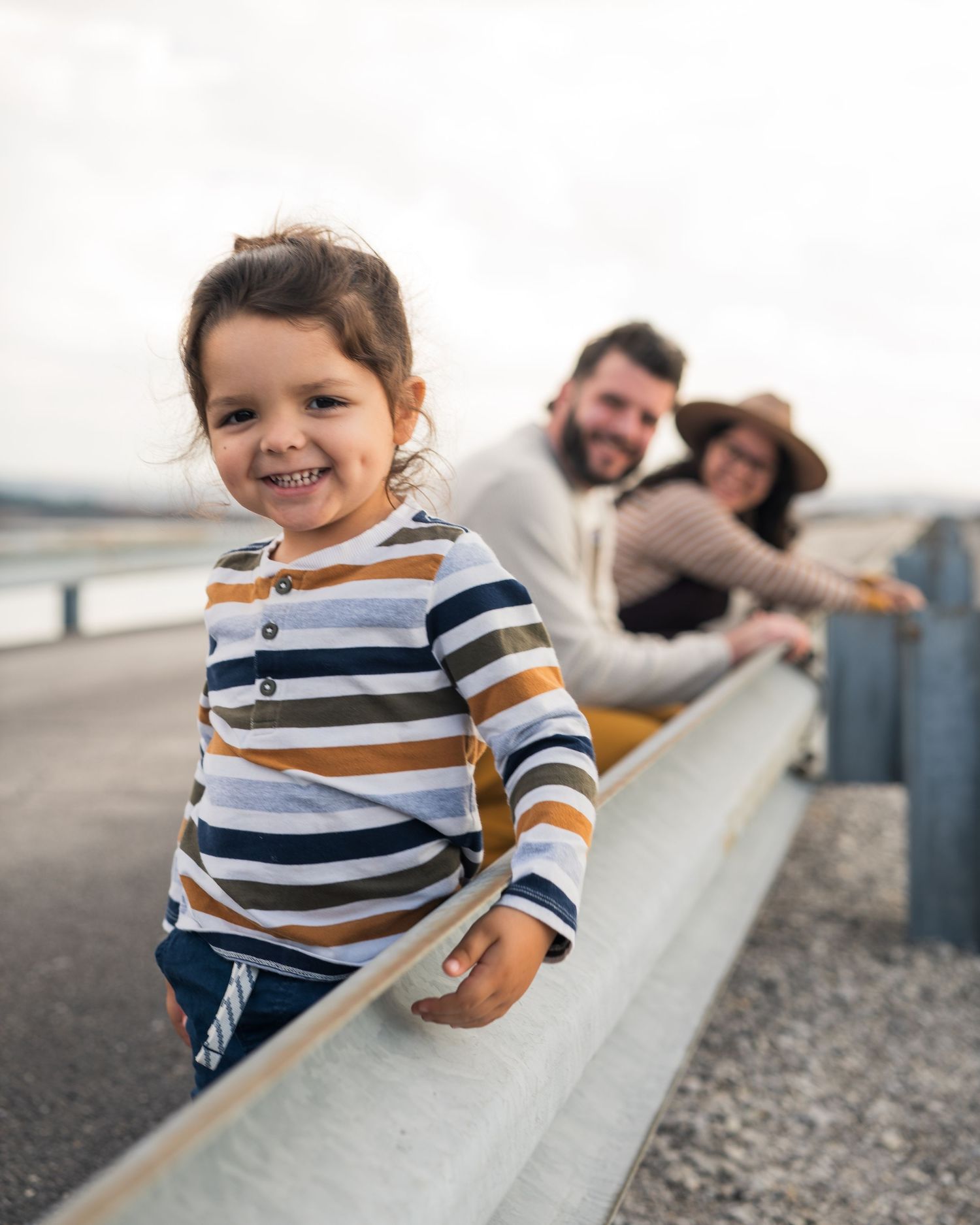 A smiling toddler in striped shirt stands by guardrail with parents smiling behind in Chattanooga, TN.