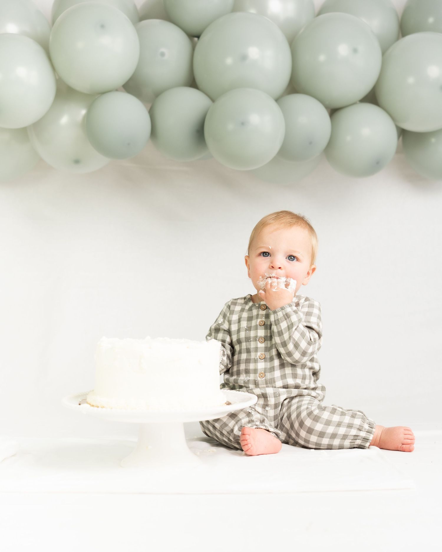 Baby in green plaid outfit enjoys first birthday cake smash with sage green balloons overhead.