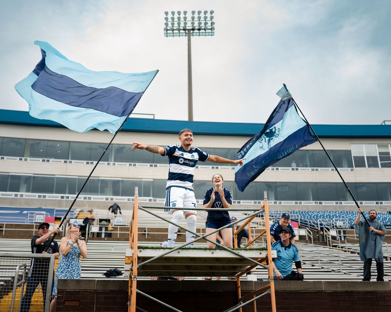 Chattanooga FC Soccer player stands triumphantly on a scaffold holding team flags at a stadium.