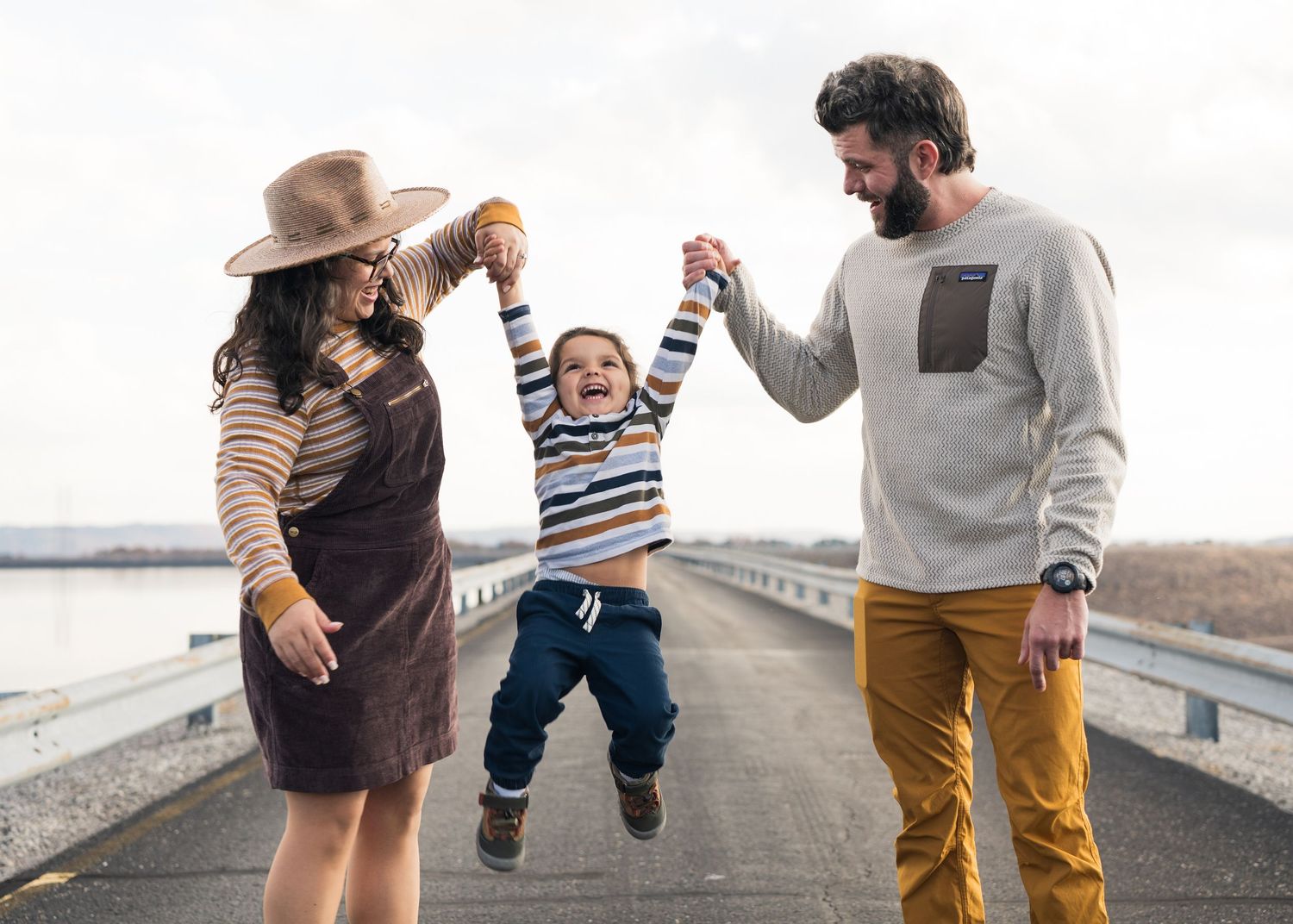 Joyful parents swing laughing toddler between them on bridge with scenic autumn landscape background in Chattanooga, TN.