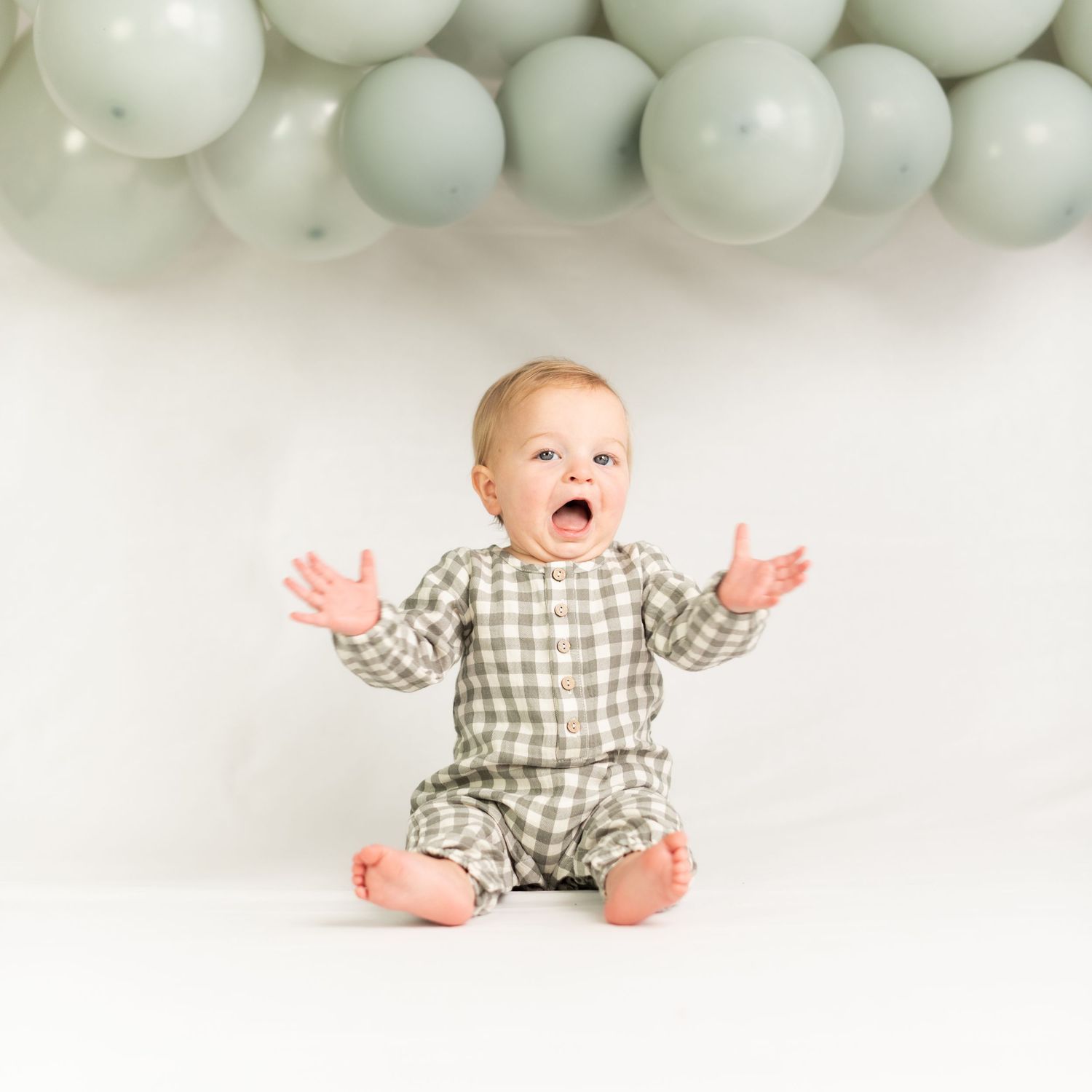 An excited baby in a plaid outfit sits with arms raised under sage green balloon decorations.
