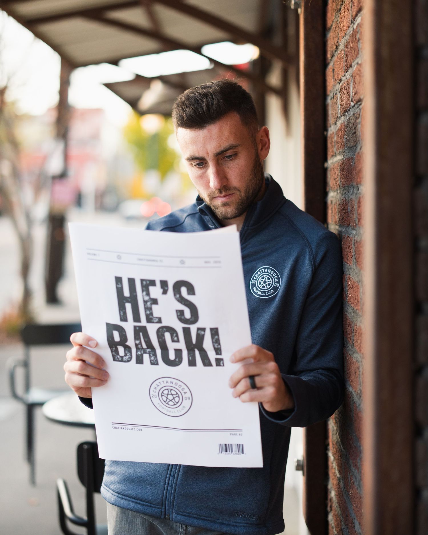Man in navy jacket reads newspaper headline HE'S BACK outside brick building. Photographed for Chattanooga FC player signing.