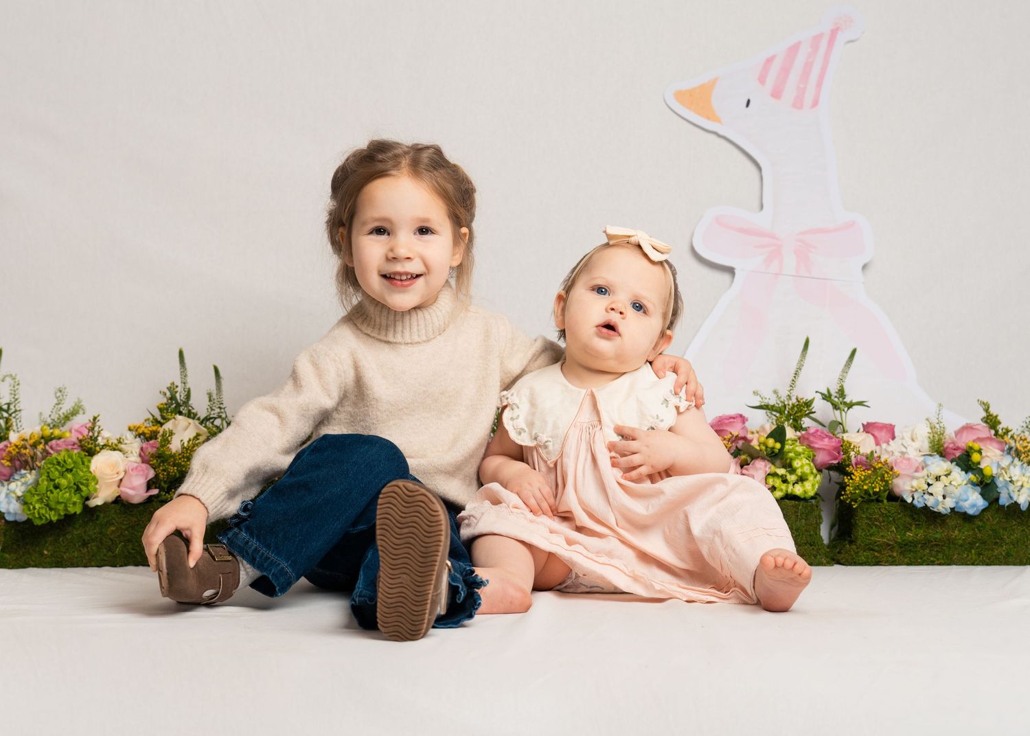 Two young sisters in spring outfits smile together in front of an Easter bunny decoration and flowers.
