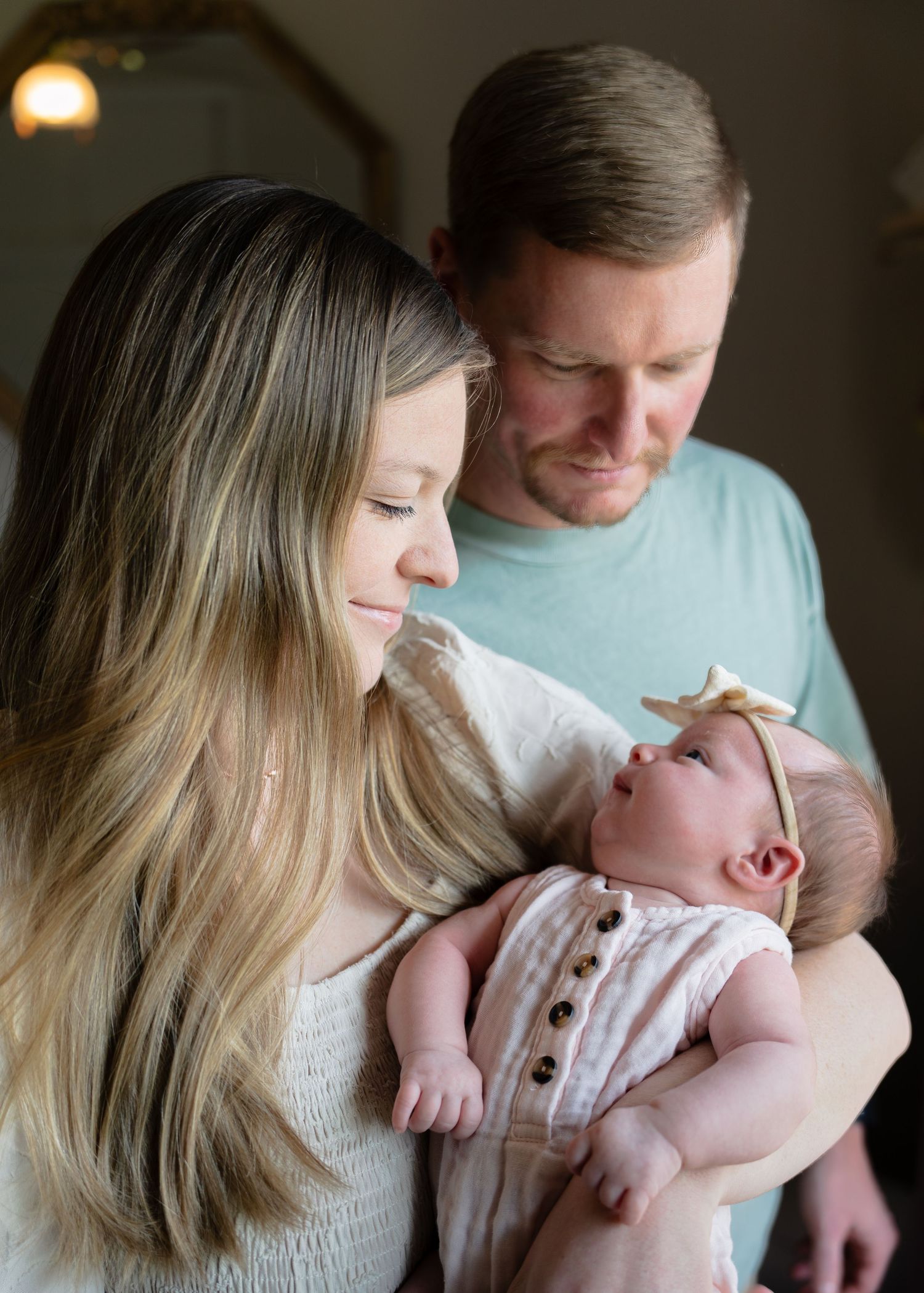 New parents tenderly gazing at newborn baby girl wearing gold bow headband.
