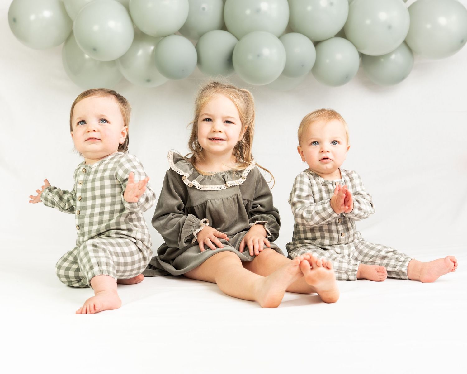 Three young children sit together in coordinating outfits with sage green balloon backdrop for birthday photos.