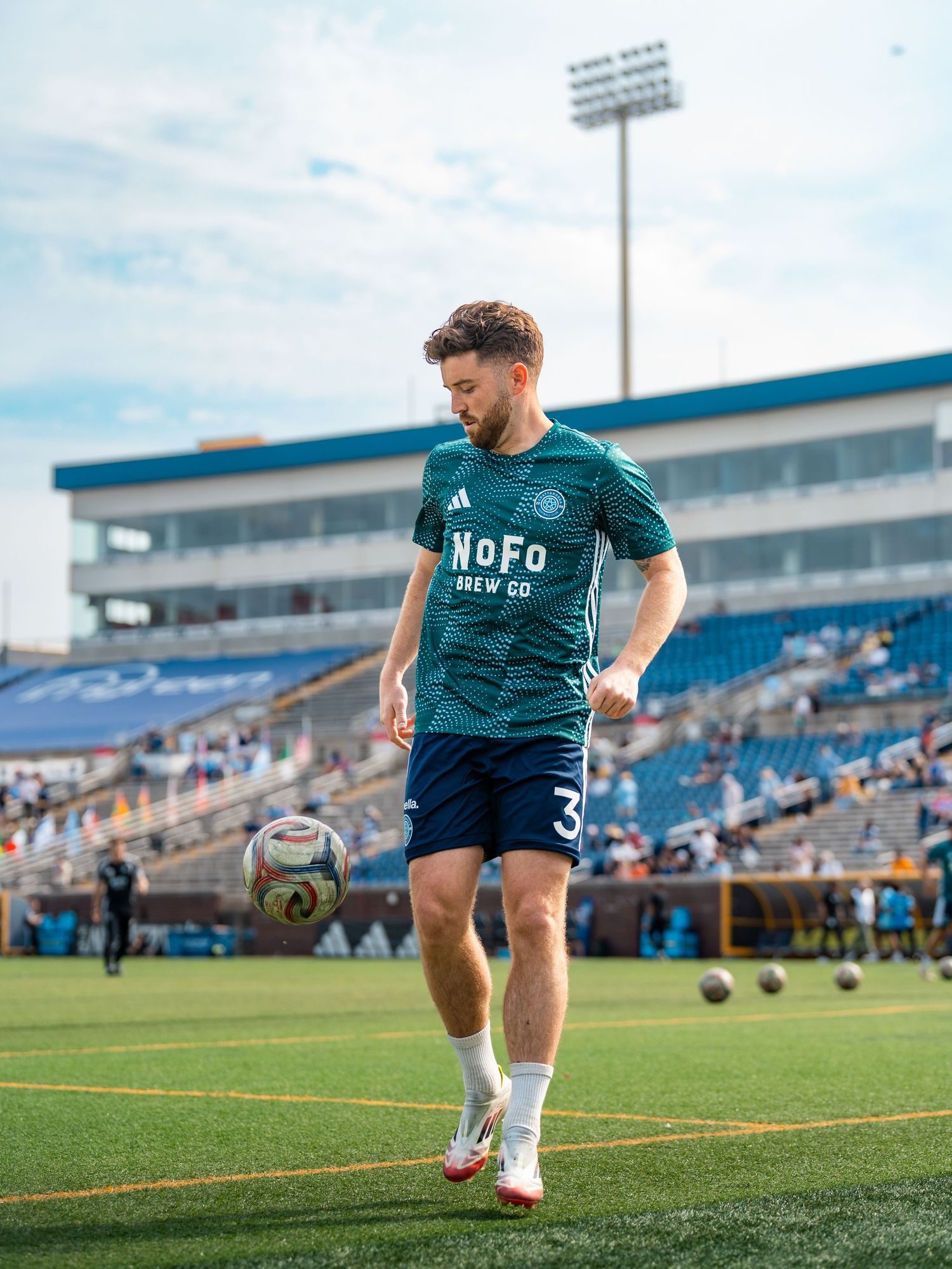 Chattanooga FC Soccer player in teal NoFo Brew Co jersey practices ball control on the field.