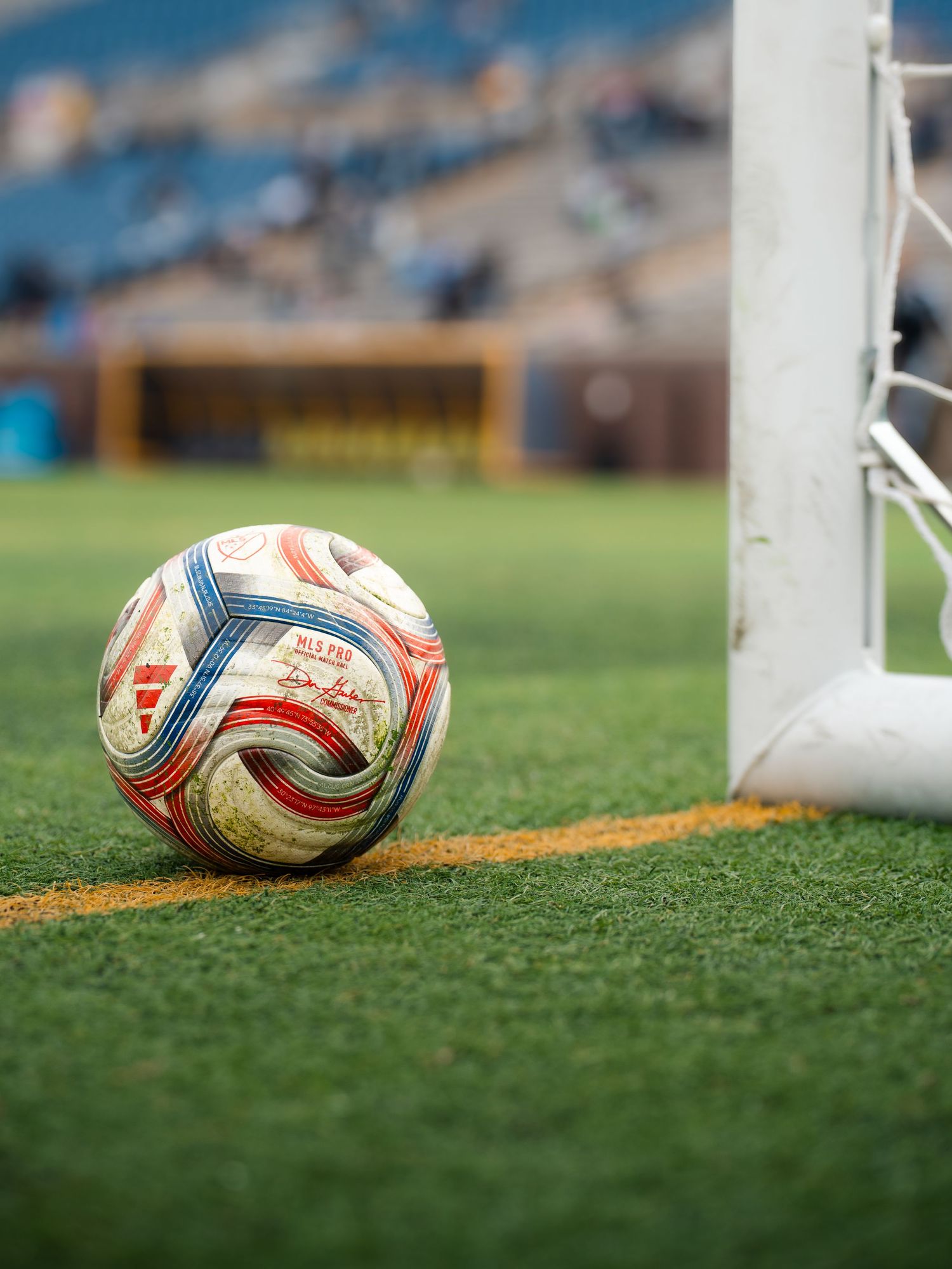 Close-up of a soccer ball resting on green turf beside a white goalpost in a stadium.