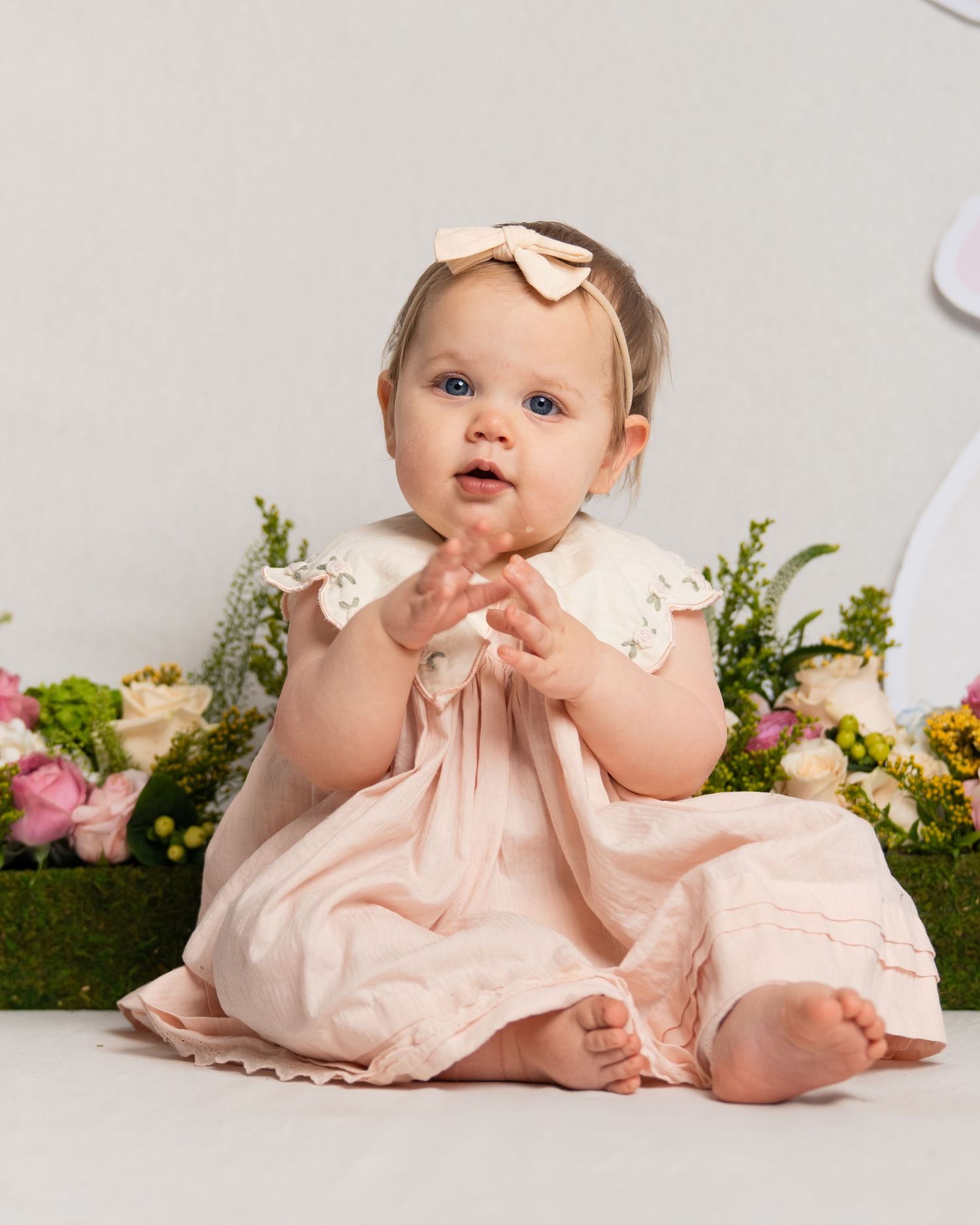 A blue-eyed baby in a pink dress with a bow headband sits among pastel spring flowers indoors.