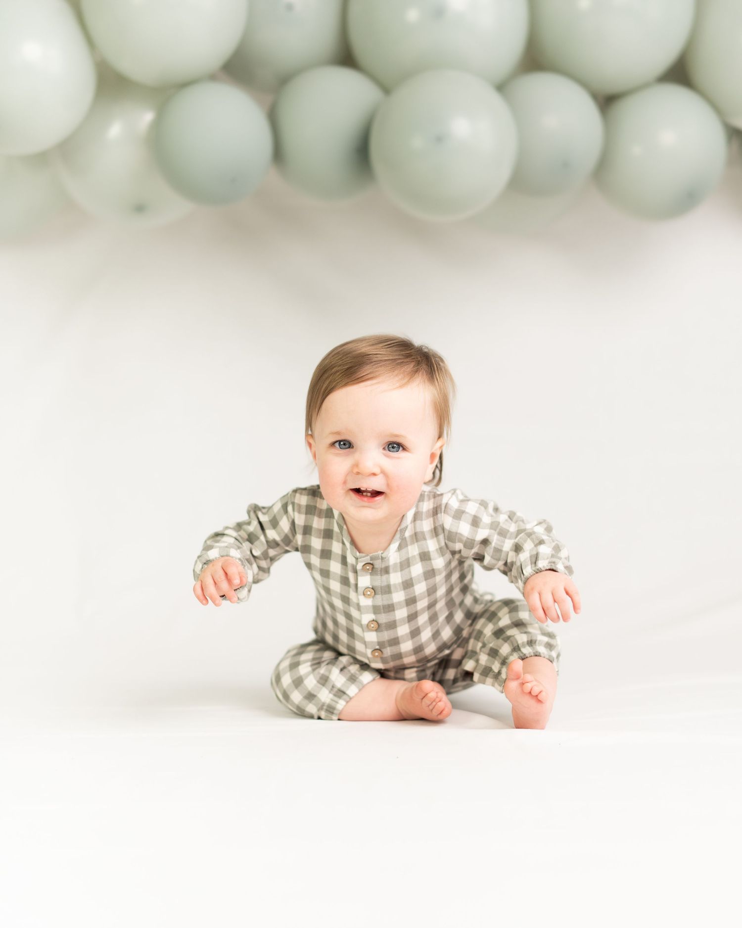 Adorable baby in sage green plaid outfit sits smiling during first birthday cake smash photo session.