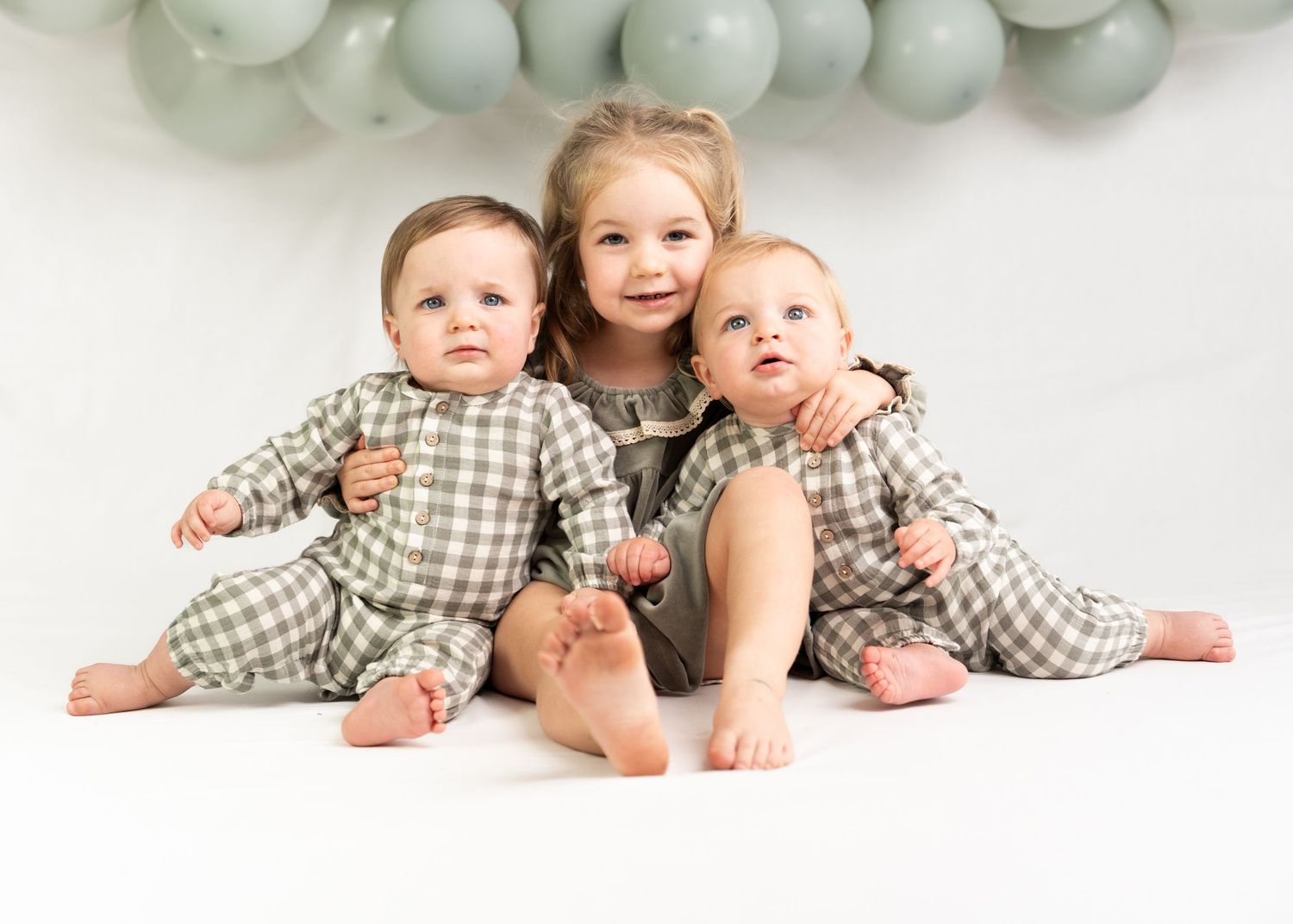 Three young siblings pose together with sage green balloons, twins in matching plaid outfits with their older sister.