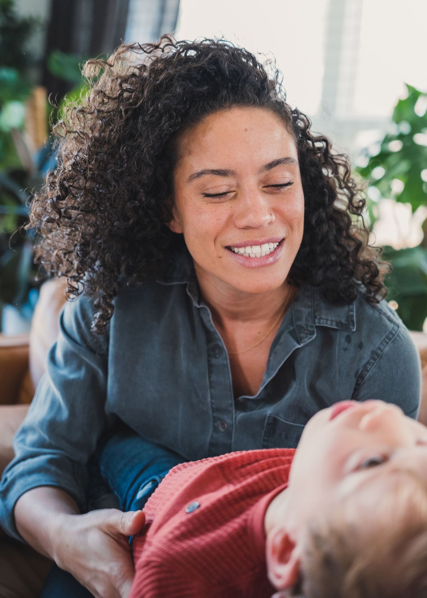 A woman smiles warmly while holding a young child indoors.
