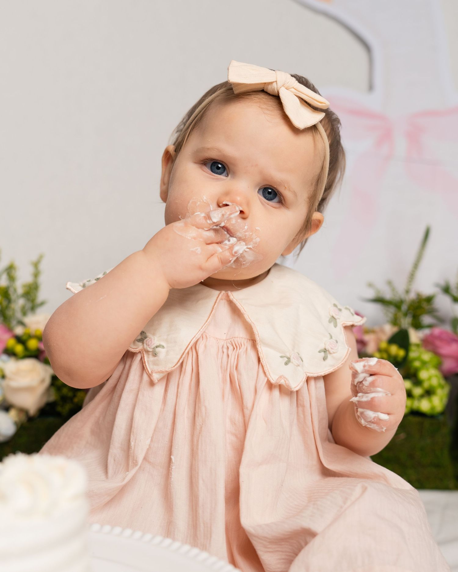 A blue-eyed baby in a pink dress tastes cake frosting with flowers and decor in the background.