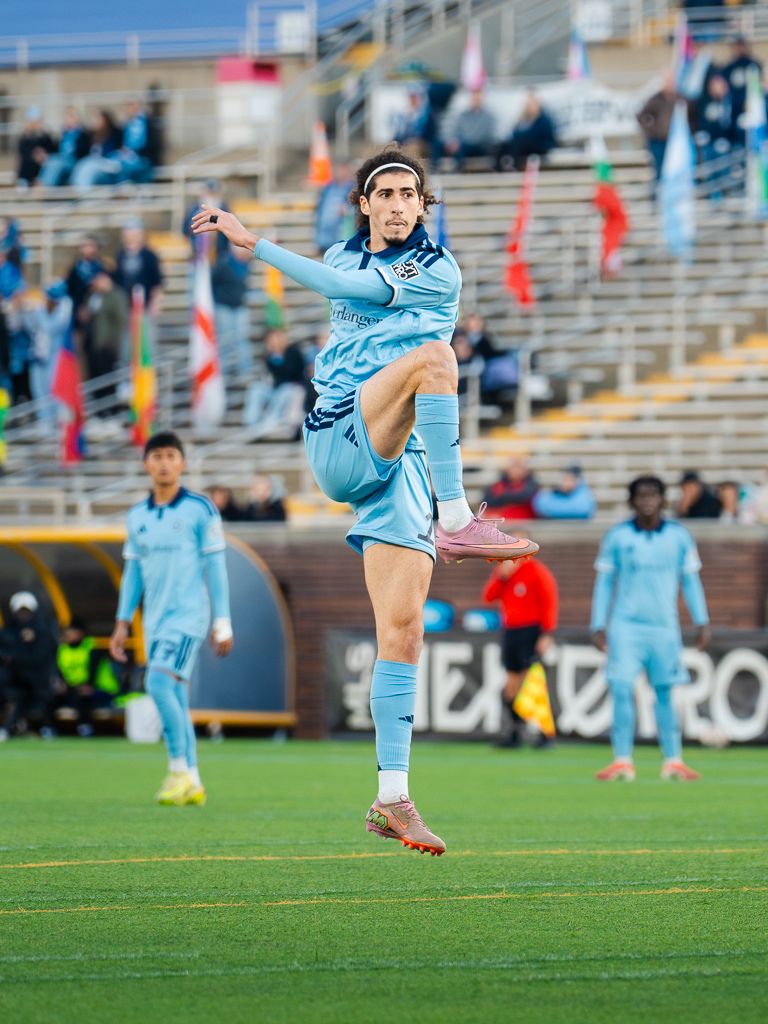 Chattanooga FC Soccer player in light blue uniform leaps mid-air during a professional soccer match in a stadium.