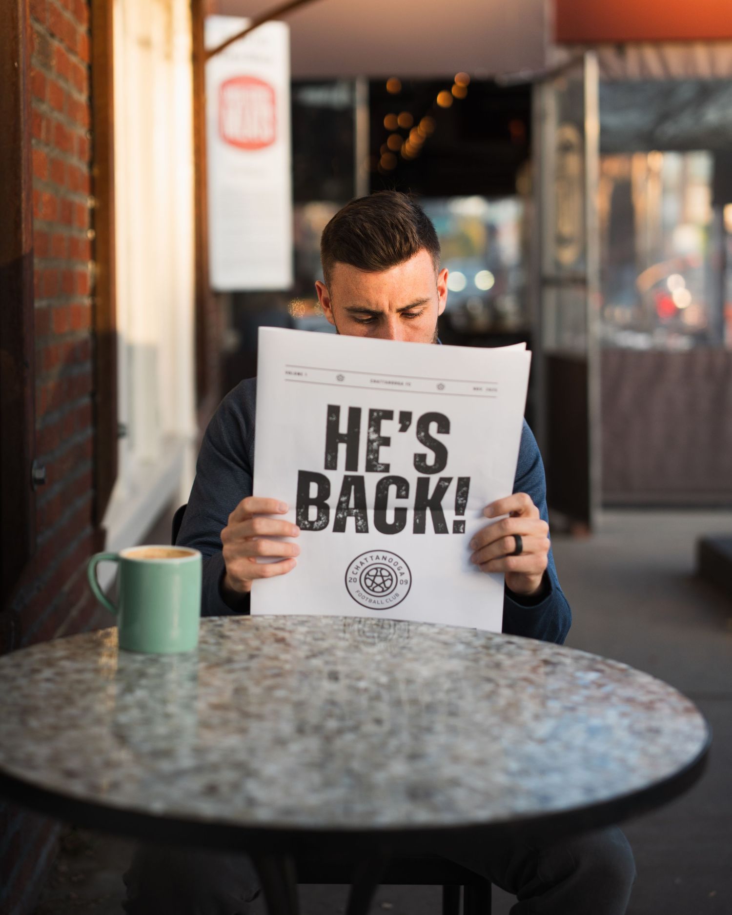 Man reads HE'S BACK newspaper headline at marble outdoor cafe table with green mug. Photographed for Chattanooga FC player signing.