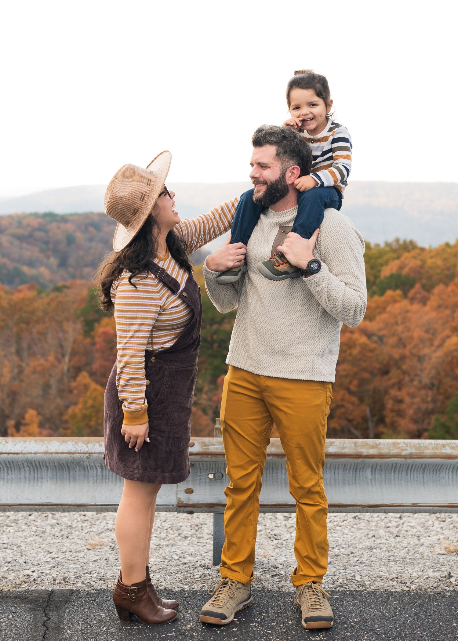 Happy family portrait with father lifting toddler on shoulders while mother smiles, fall foliage backdrop in Chattanooga, TN.