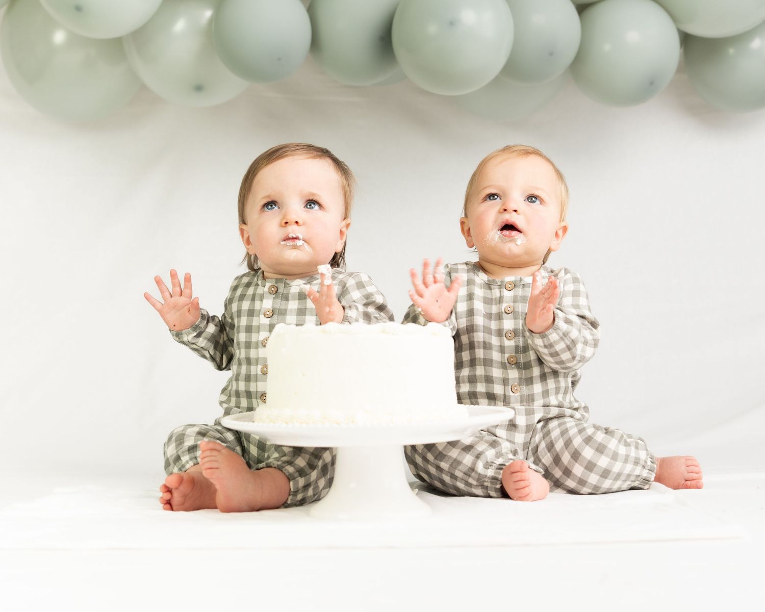 Twin babies in matching plaid outfits sit with white birthday cake under sage green balloon arch.