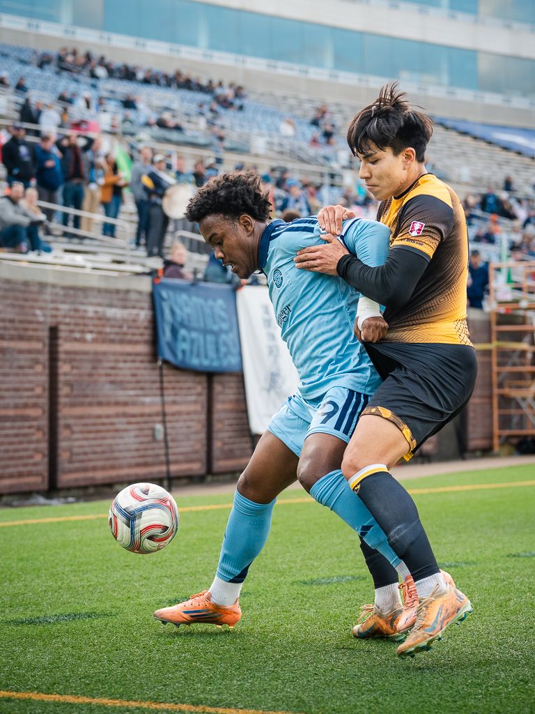 Two soccer players battle for possession during a competitive match, one in light blue and one in gold.