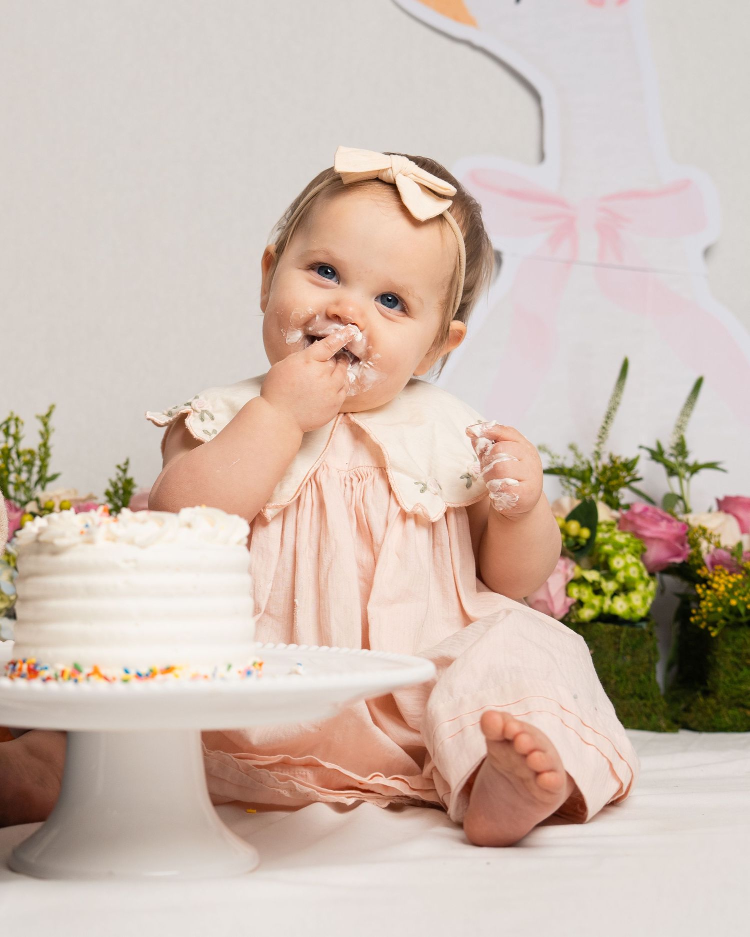 A joyful baby in a pink dress digs into a white frosted birthday cake with sprinkles.