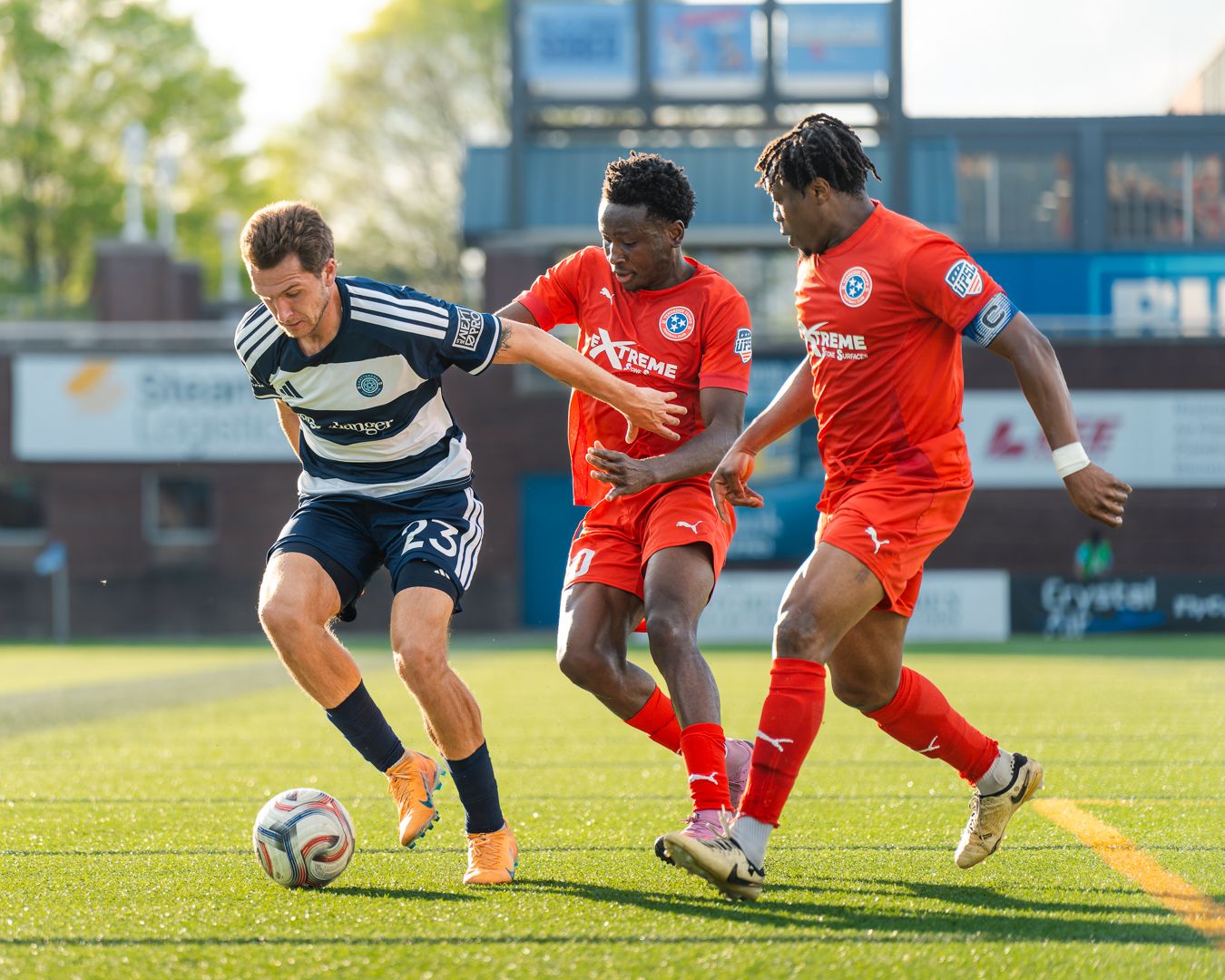 Two soccer players in red jerseys compete for the ball against a player in a navy blue uniform.