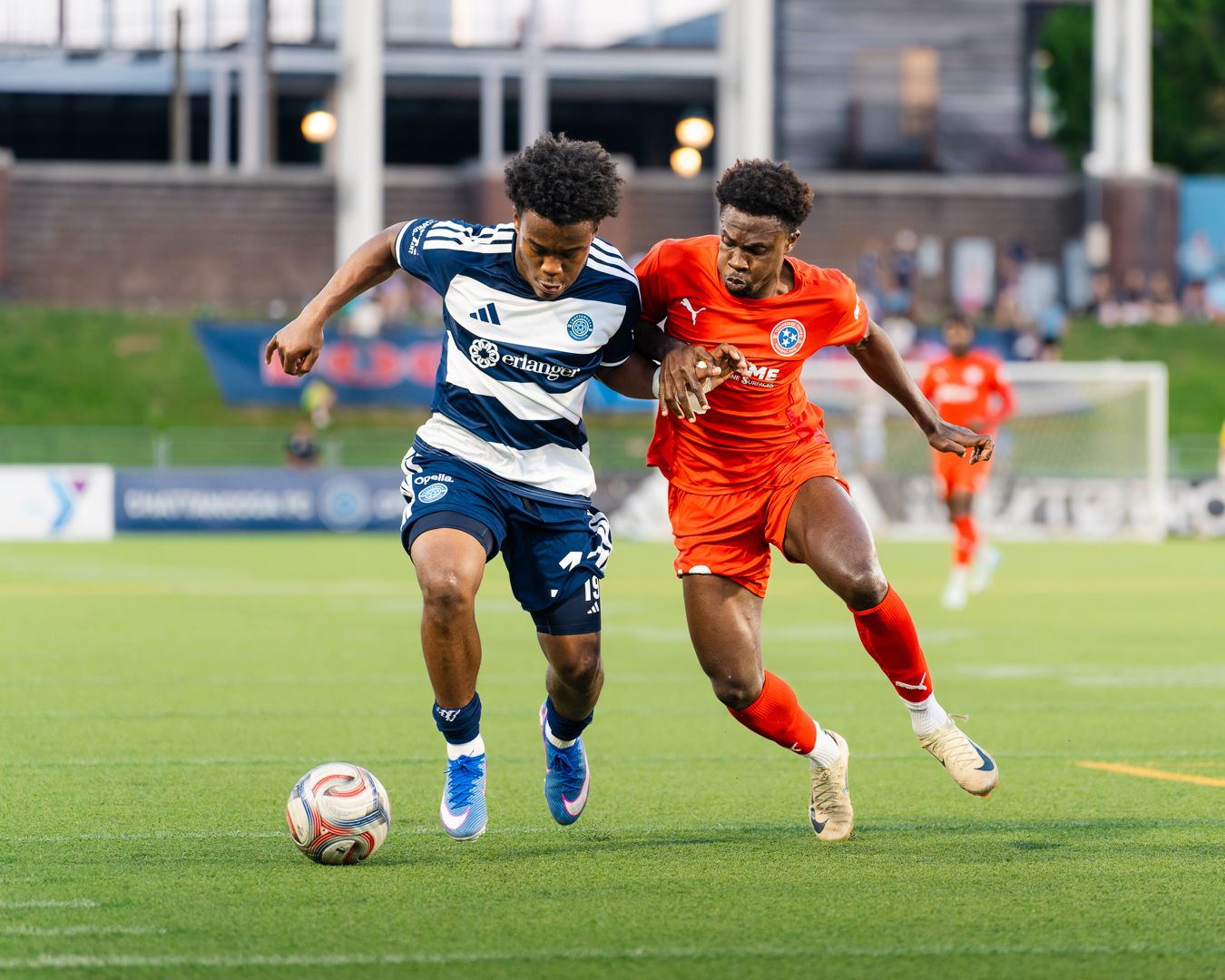 Two soccer players battle for possession during a competitive U.S. Open Cup match, one in navy blue and one in orange.