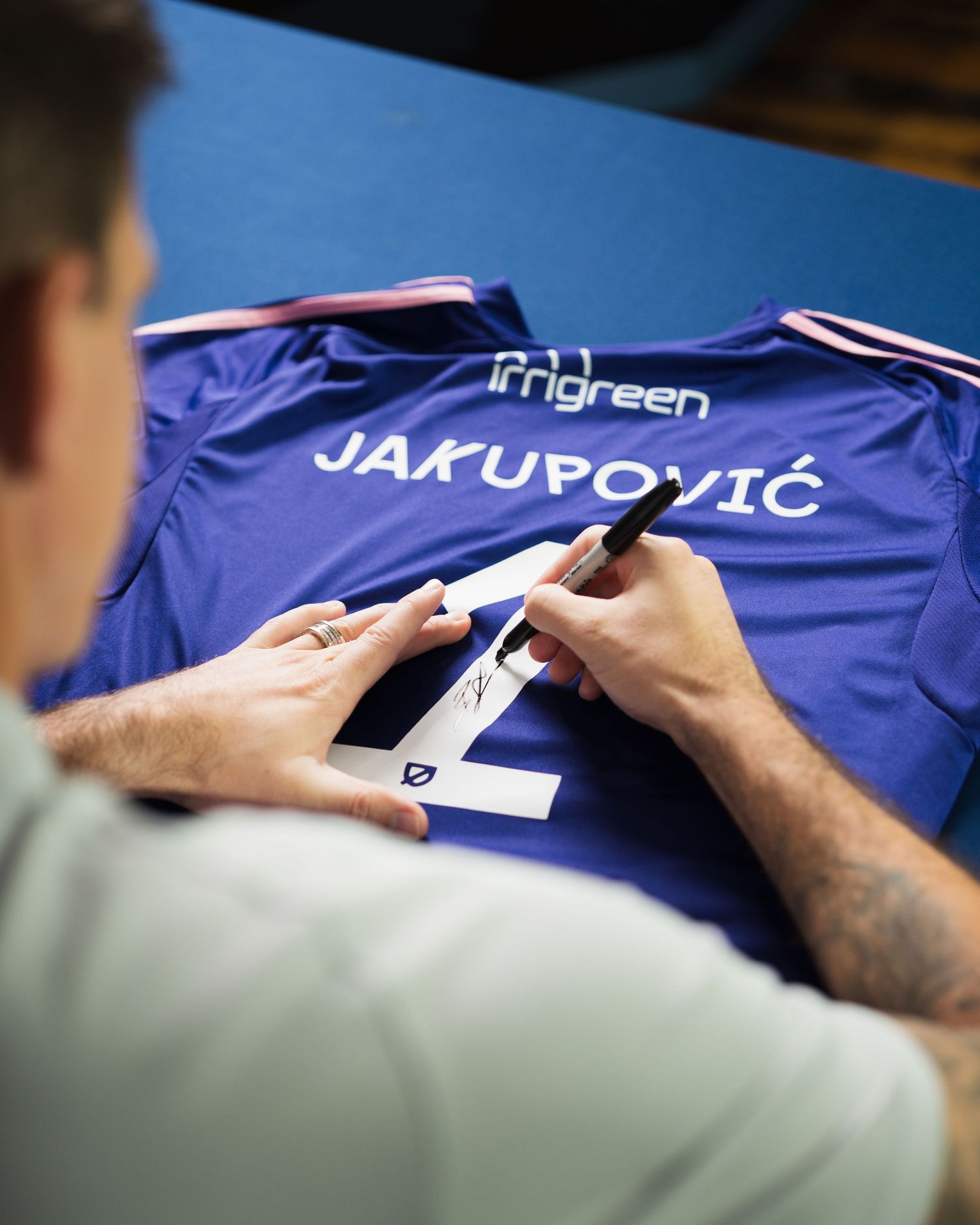 Soccer player signs the back of a purple Jakupovic jersey on a blue table. Photographed for Chattanooga FC player signing.