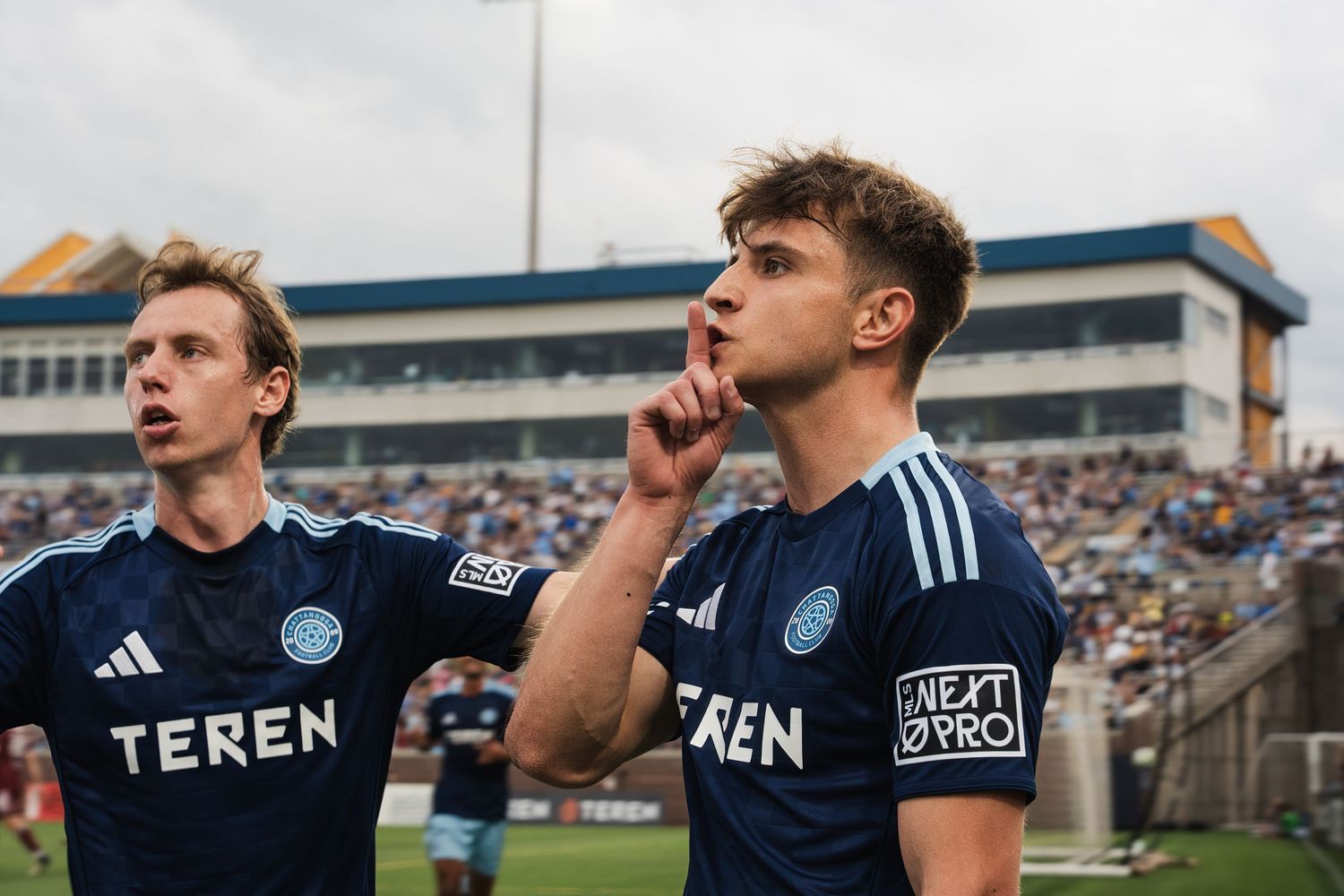 Two Chattanooga FC soccer players in dark navy Teren jerseys celebrate goal with silence gesture.
