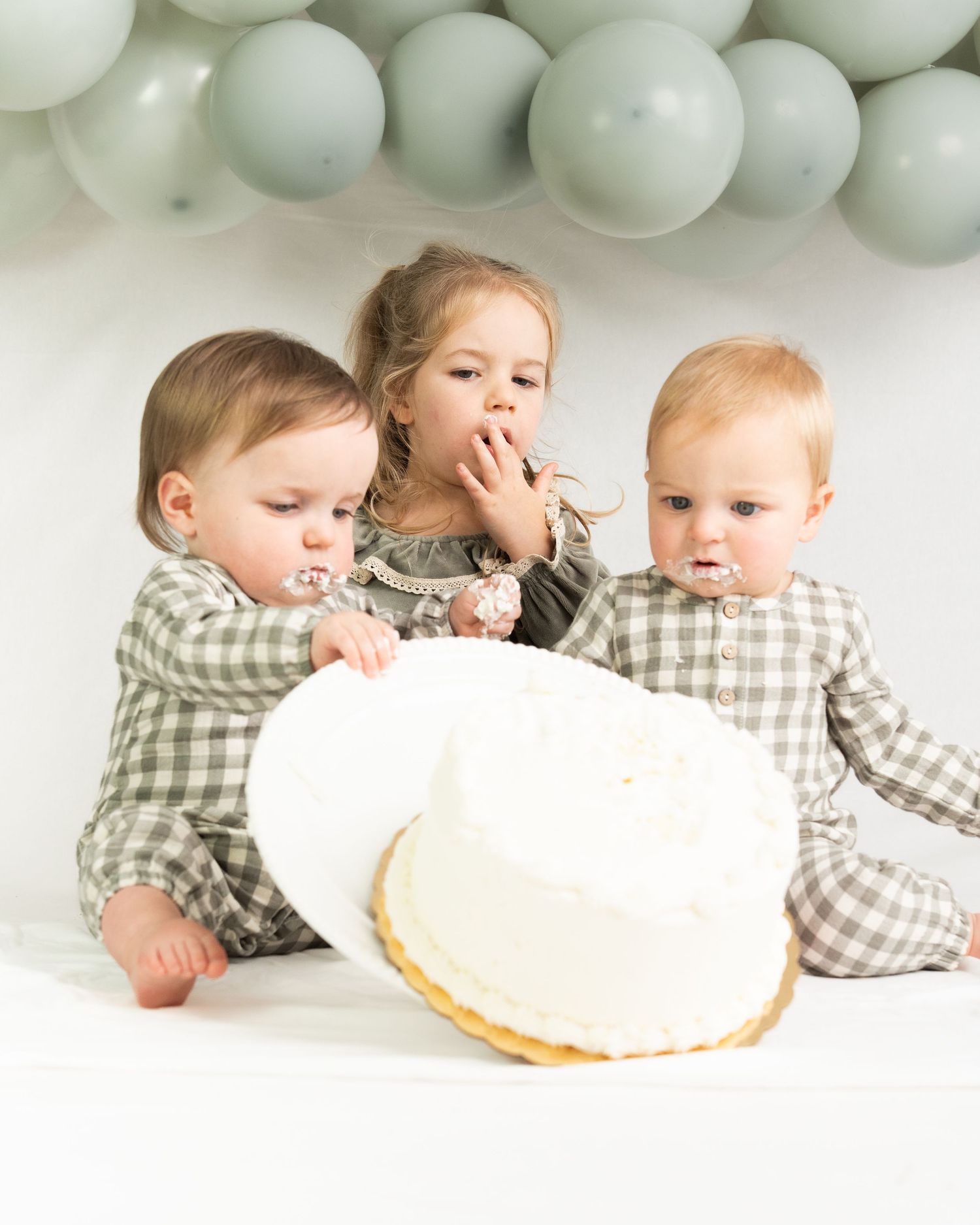 Three young siblings gather around a white birthday cake with sage green balloon garland backdrop.