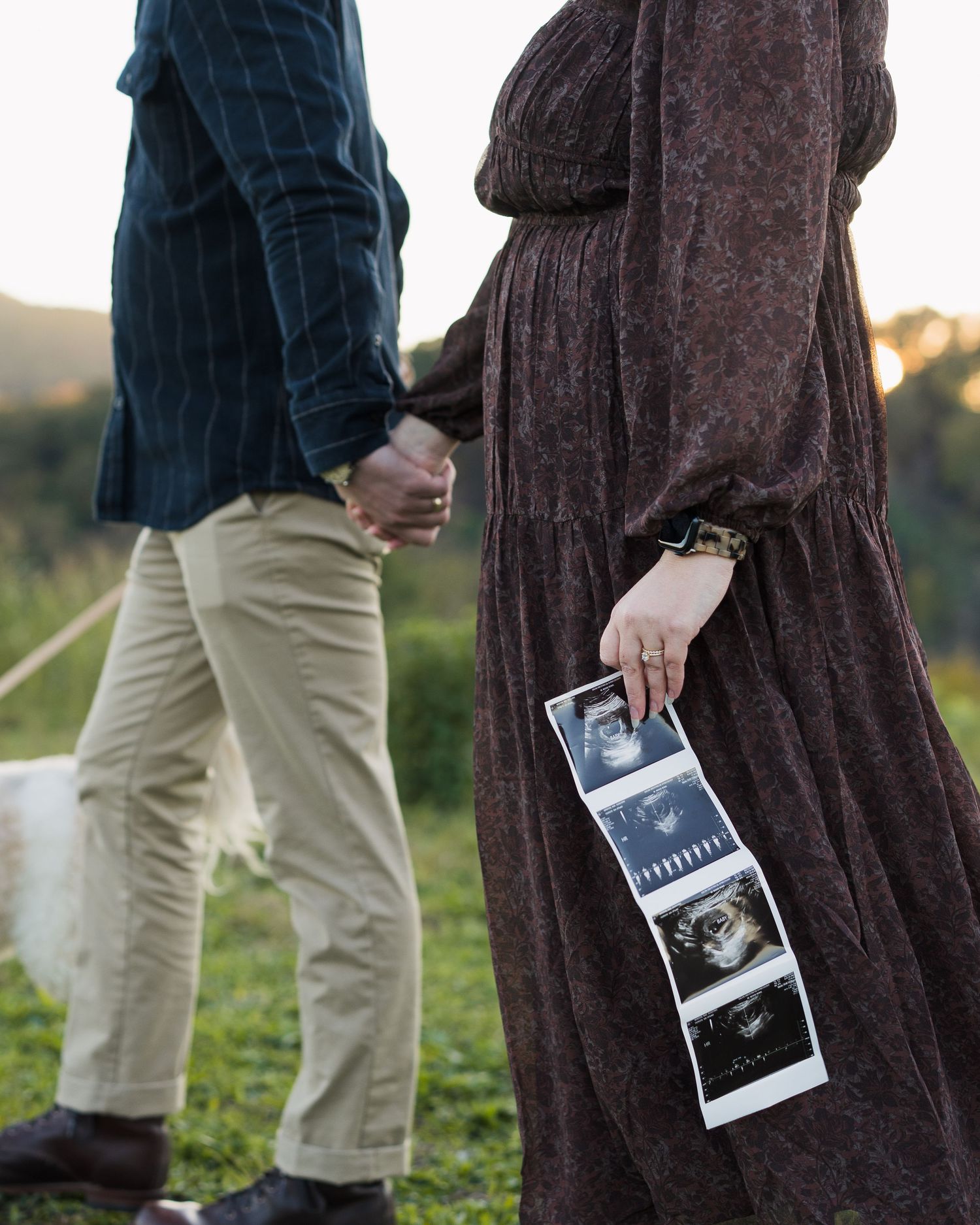 Pregnant woman holds ultrasound photos while couple walks hand-in-hand in green field in Chattanooga, TN.