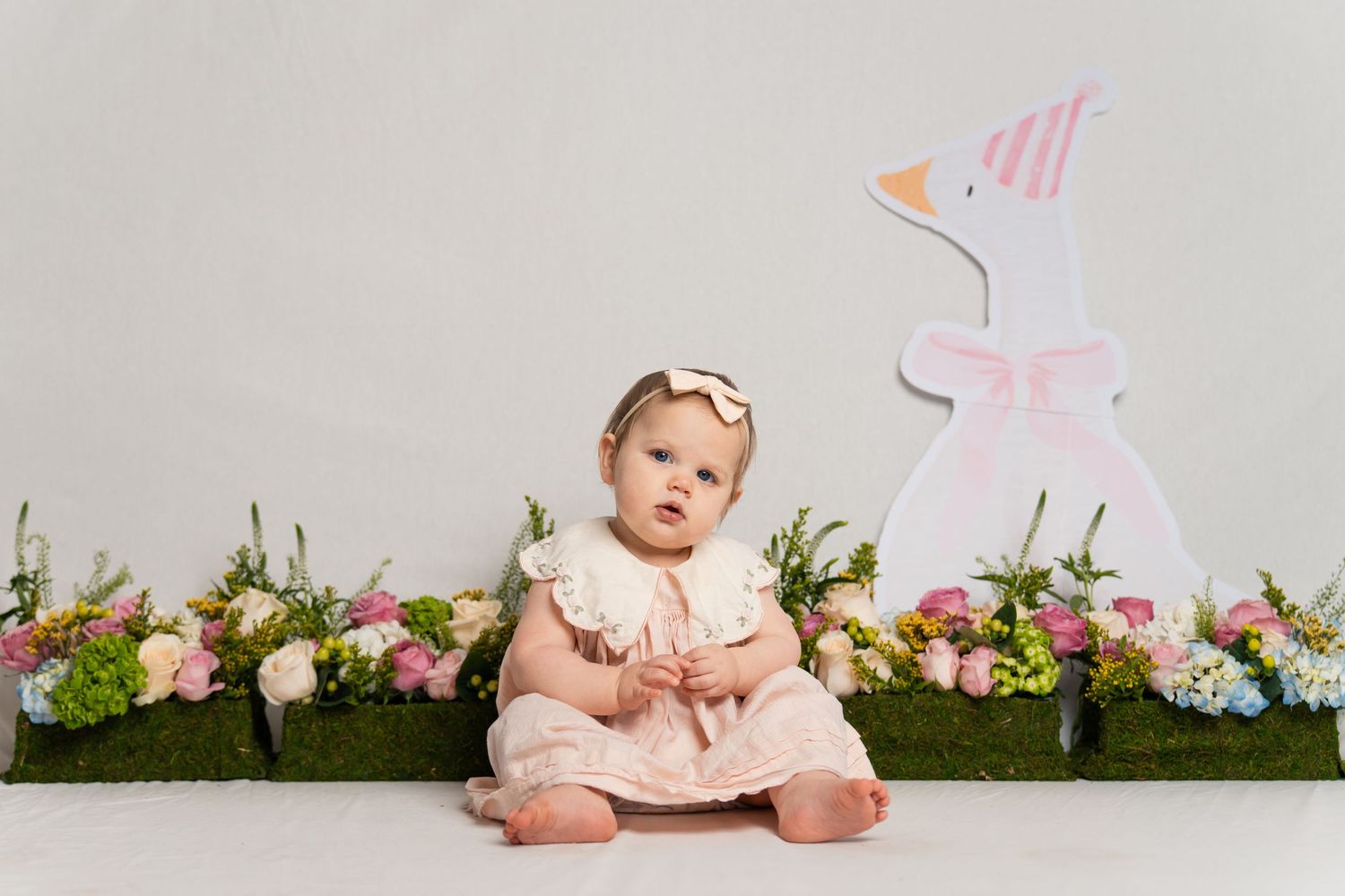An adorable baby in a pink dress sits before a decorative Easter bunny and colorful spring flowers.