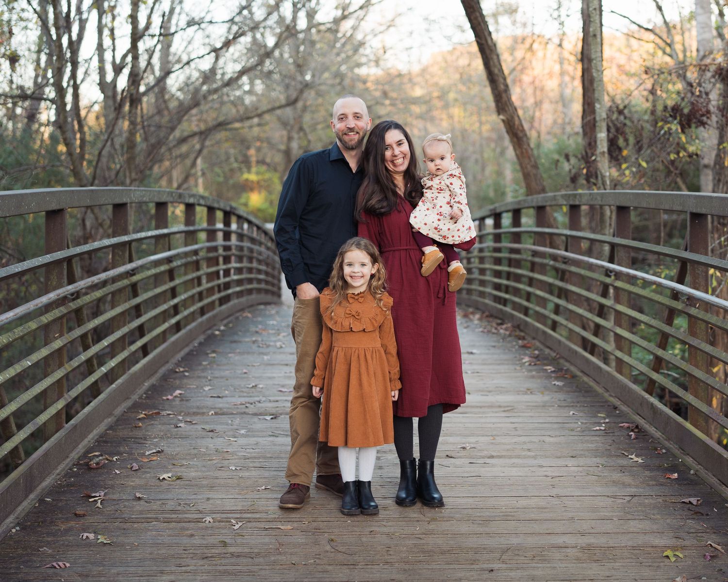Family of four poses on wooden bridge surrounded by autumn trees in fall portrait in Chattanooga, TN.