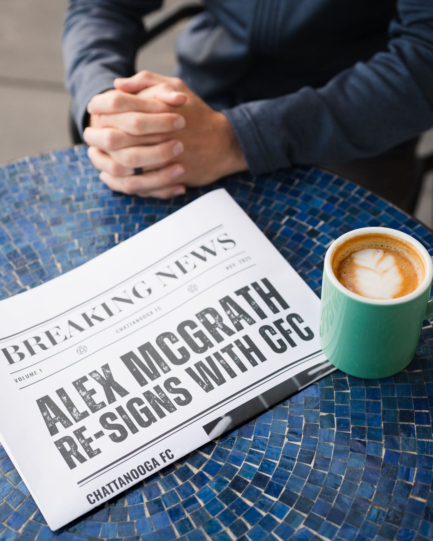 Breaking news newspaper reading Alex McGrath Re-Signs with CFC beside a latte on a mosaic table. For Chattanooga FC player signing.