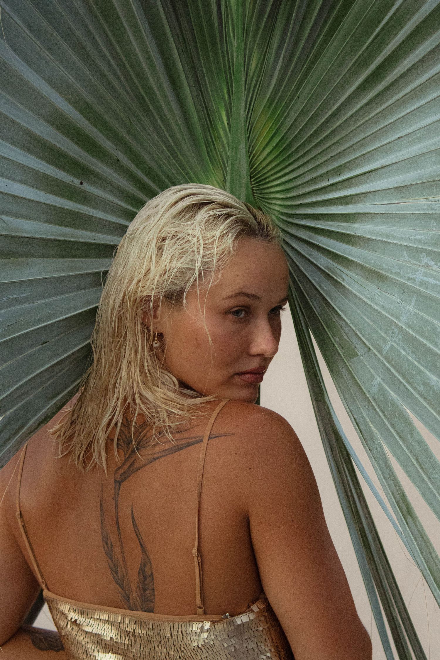 Blonde woman with shoulder tattoo in gold sequin top posing beside large green tropical palm leaf.
