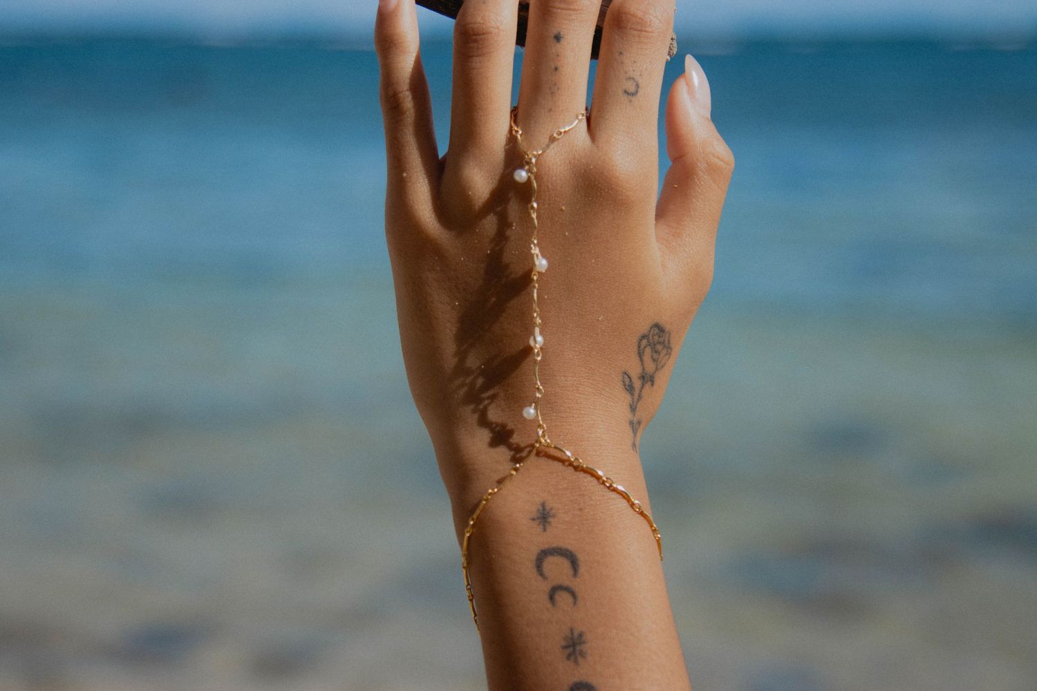 Close-up of a hand with tattoos wearing a delicate gold chain hand bracelet against a blurred ocean background.
