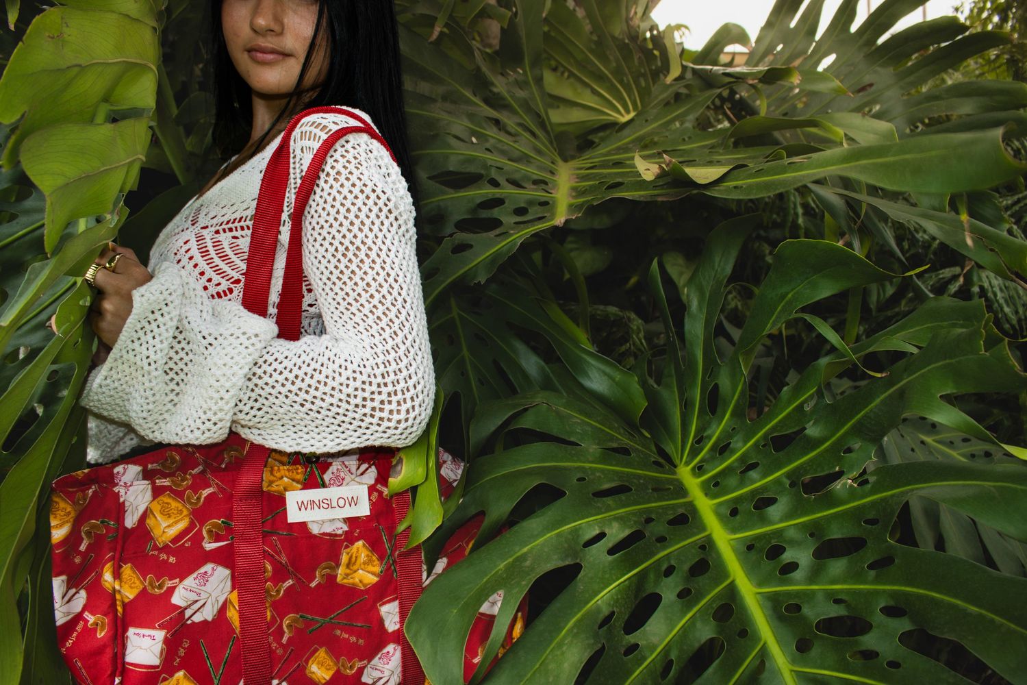 Woman in white crochet sweater holding a red Winslow tote bag with colorful print among tropical leaves.