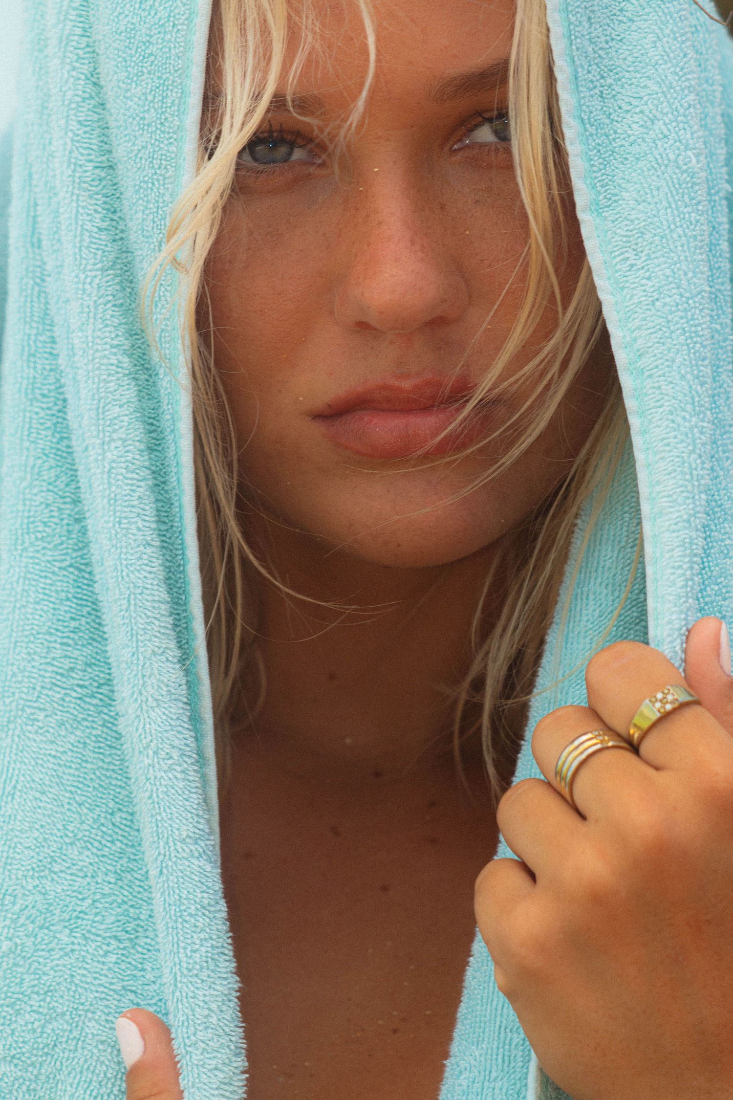 Blonde woman with blue eyes wrapped in teal towel wearing stacked rings on sandy beach.