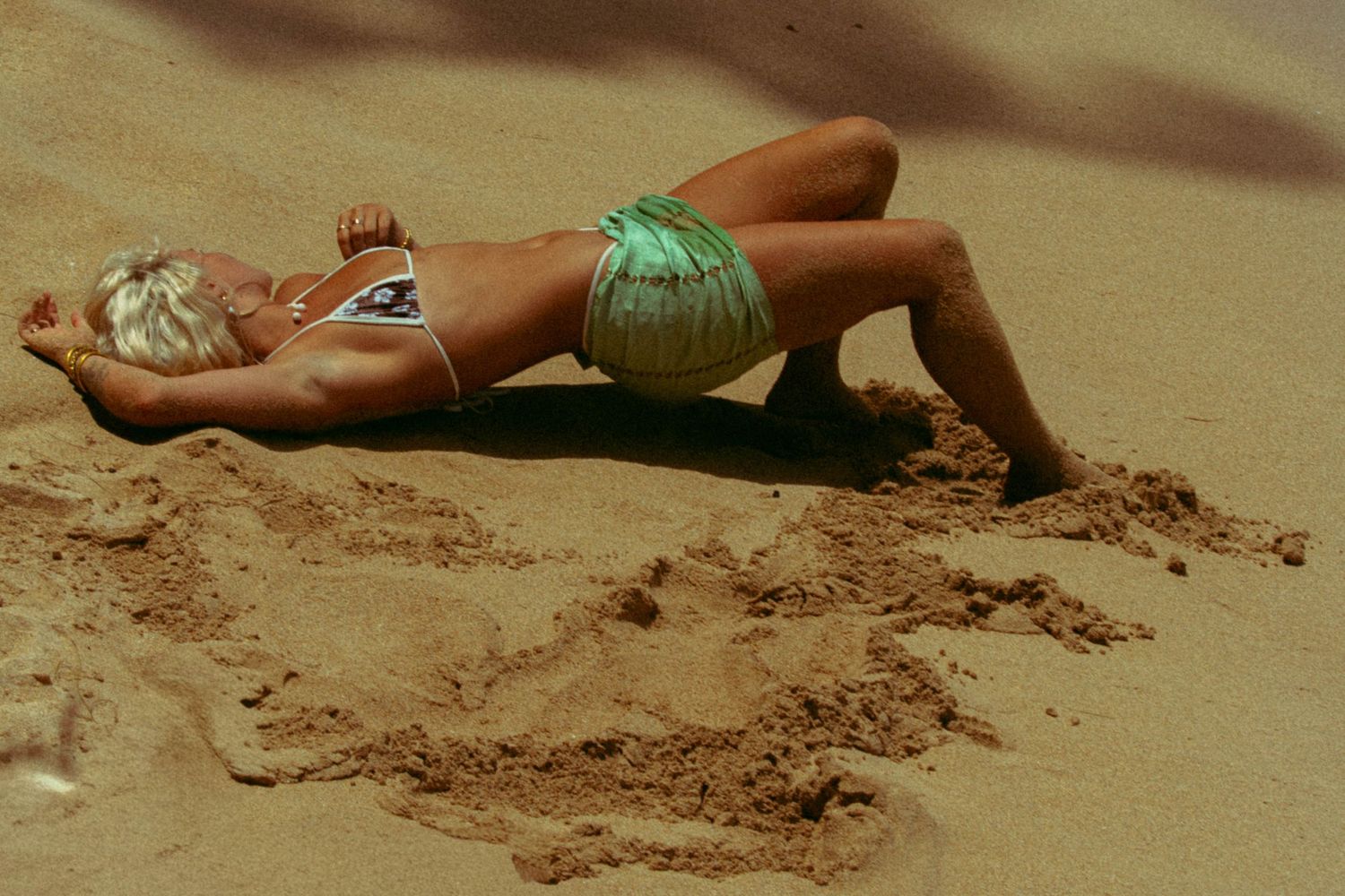 Blonde woman sunbathing in green bikini bottoms lying face down on warm sandy beach.