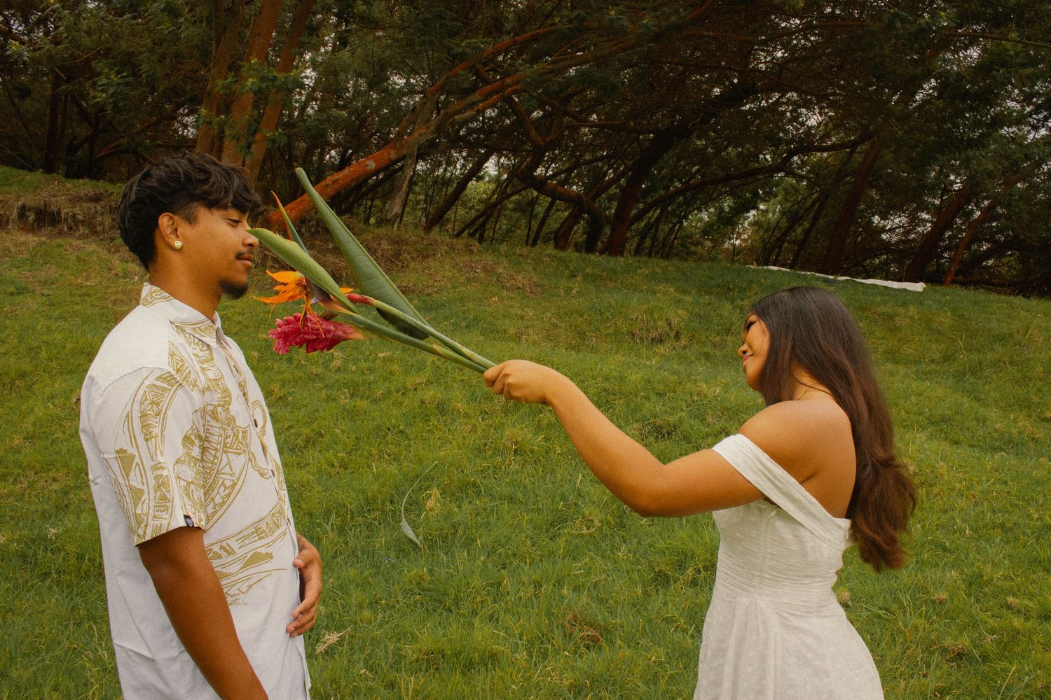 Young woman in a white dress offering tropical flowers to a man on a green grassy hillside.