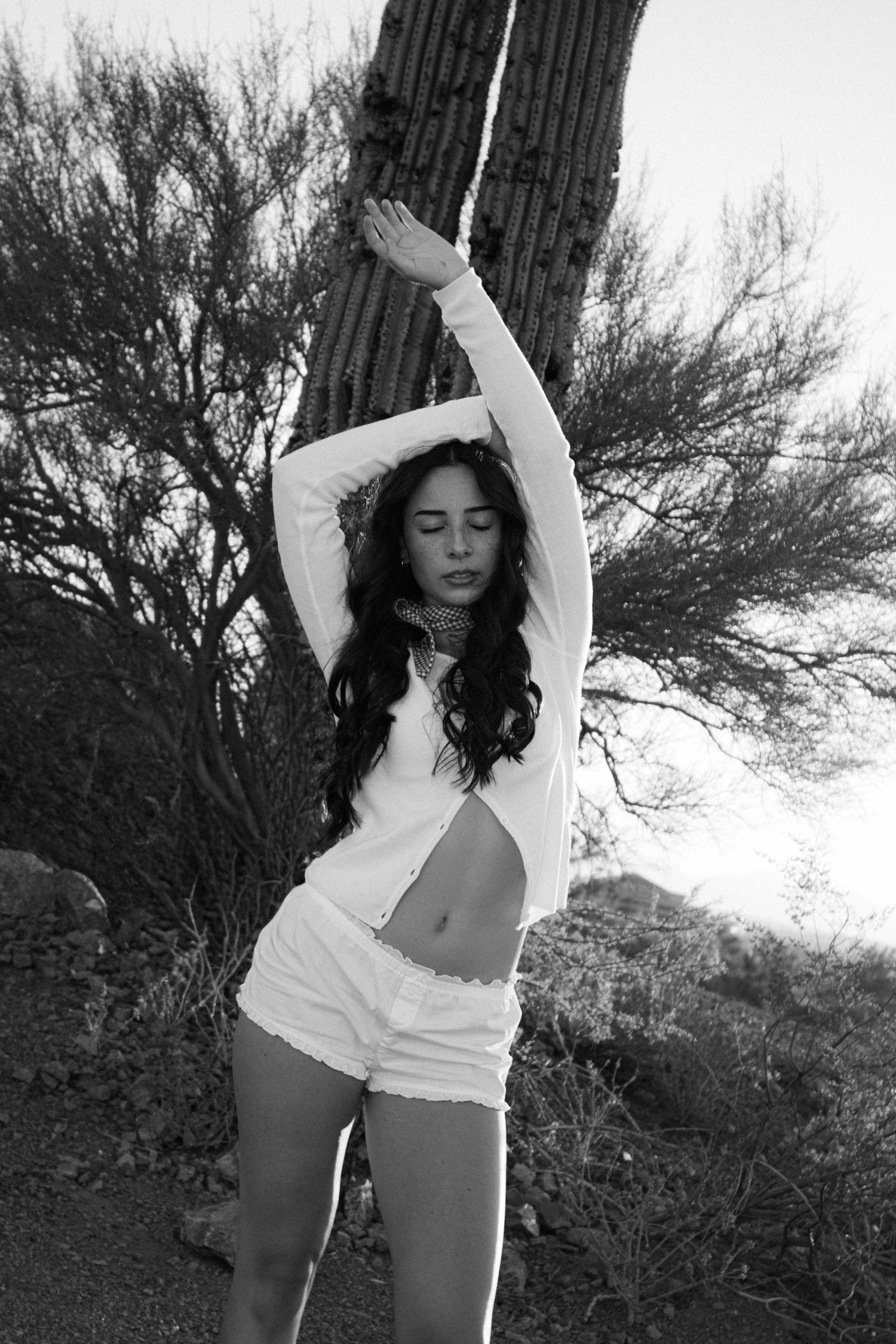 Black and white photo of woman stretching arms up beside saguaro cactus in desert.