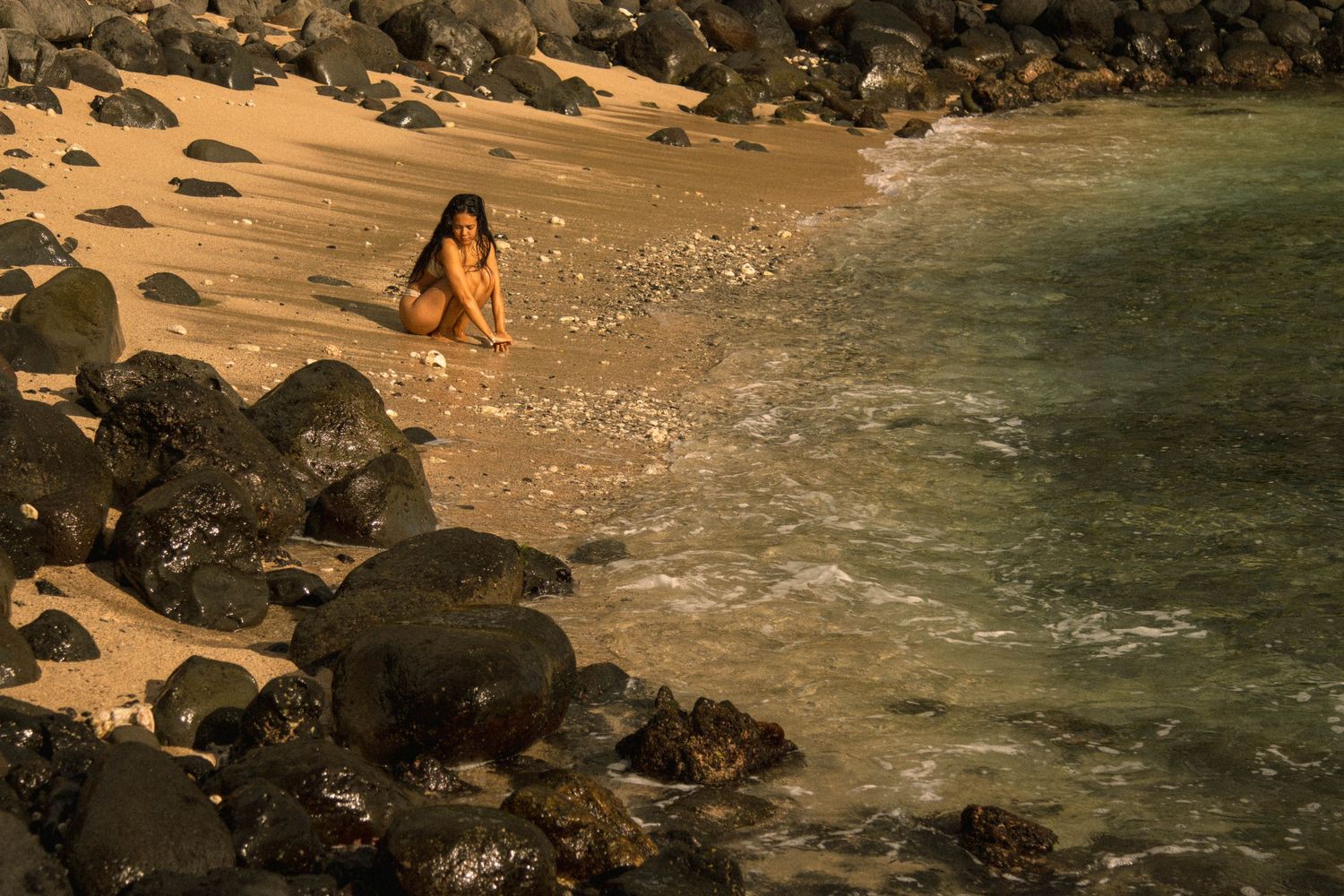 Woman sitting alone on sandy beach surrounded by dark volcanic rocks at sunset.