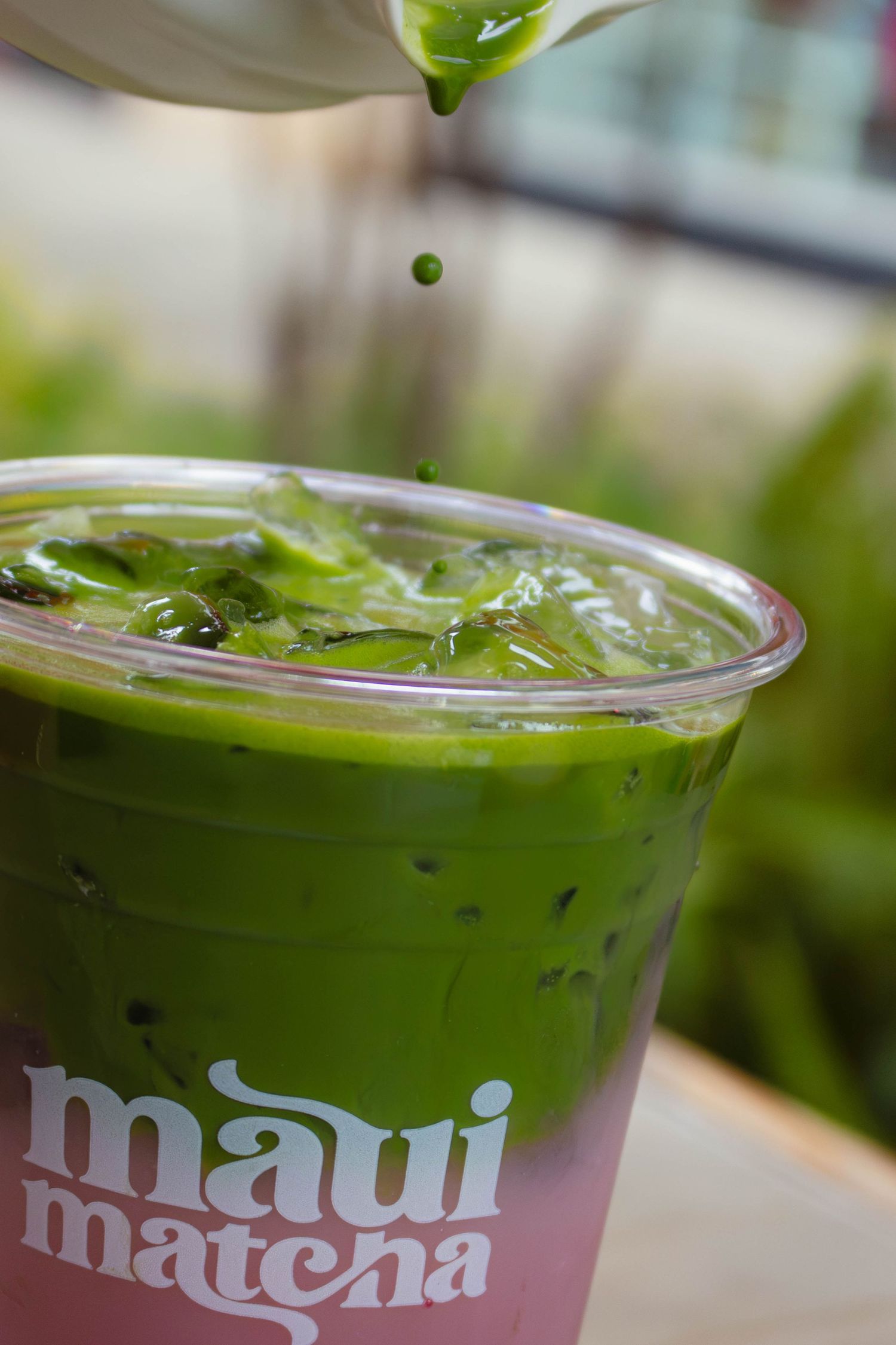 Vibrant green matcha being poured into an iced drink in a pink Maui Matcha cup outdoors.