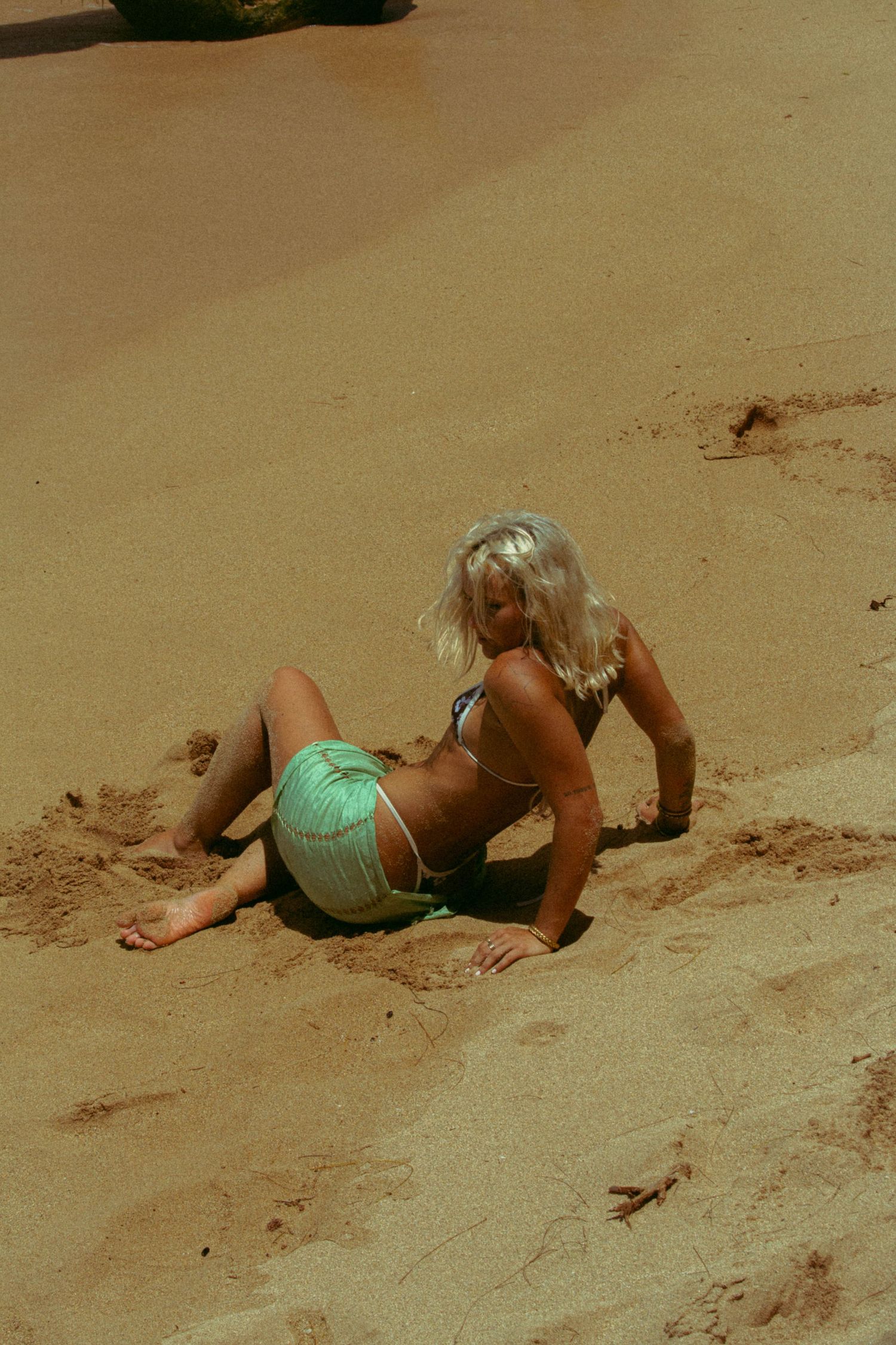 Blonde woman in green bikini sitting on sandy beach posing in bright sunlight.