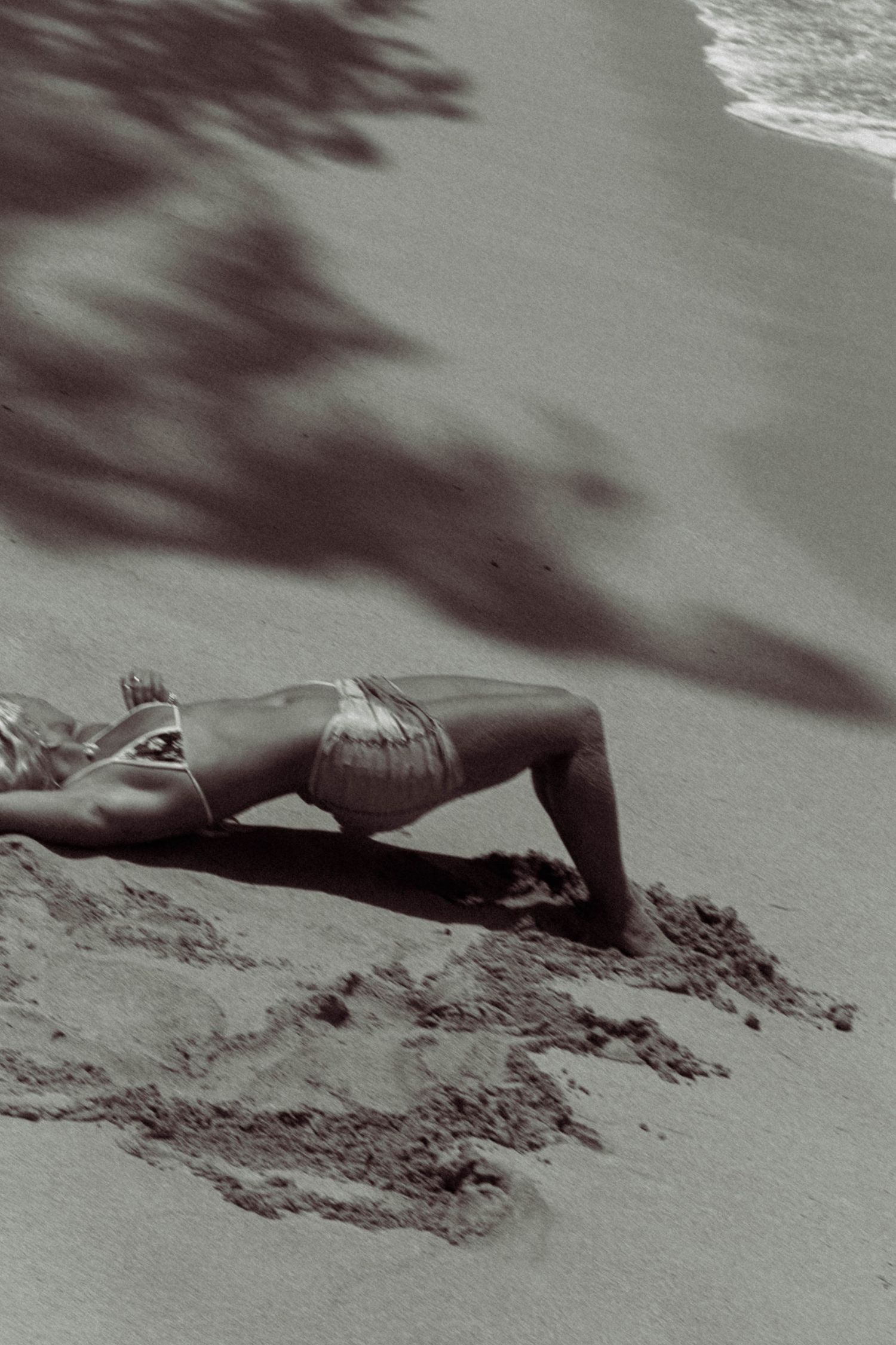 Black and white aerial view of woman in bikini lying on sandy beach near ocean waves.